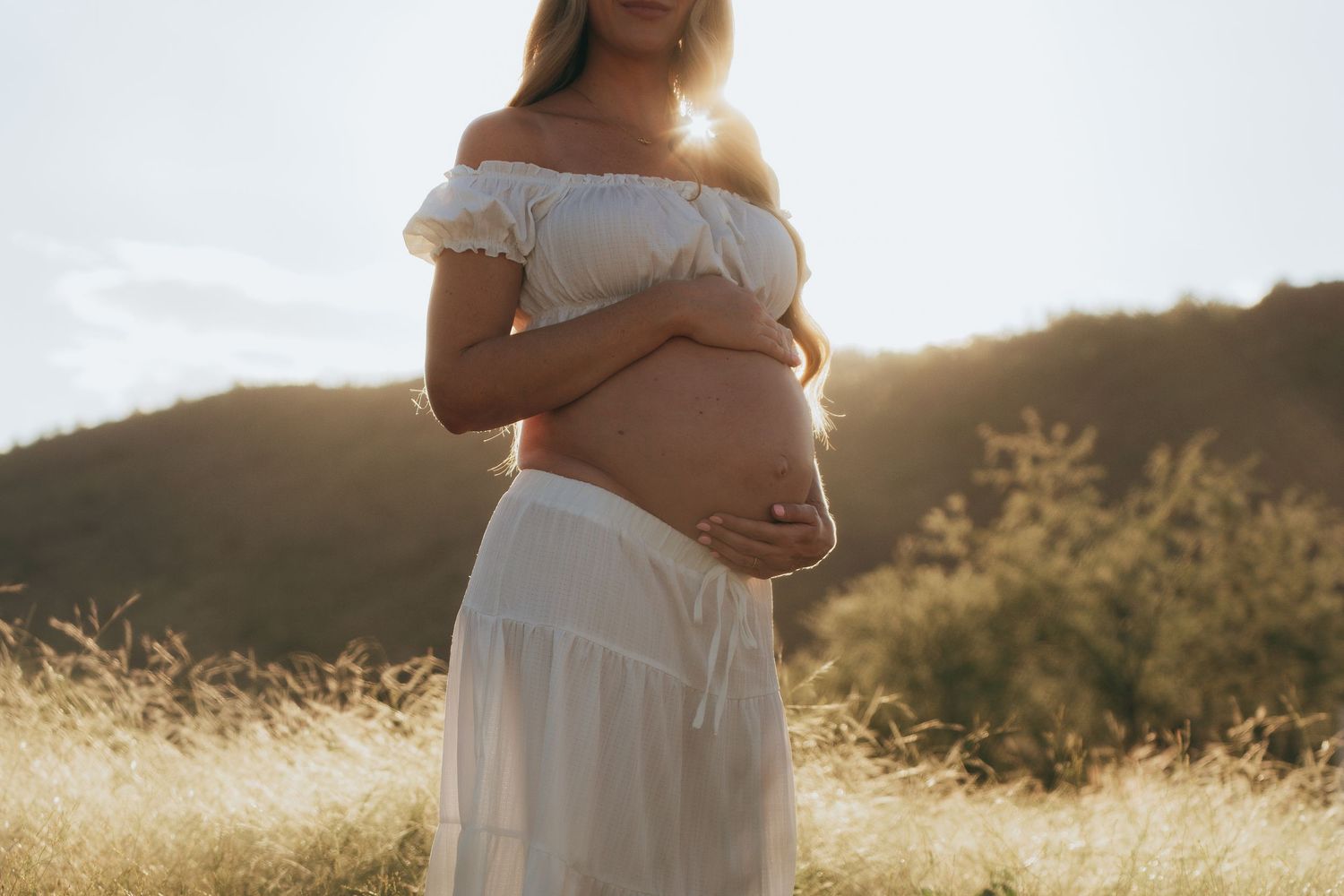 Pregnant woman in white off-shoulder dress cradling baby bump in golden sunlit field with hills in background.