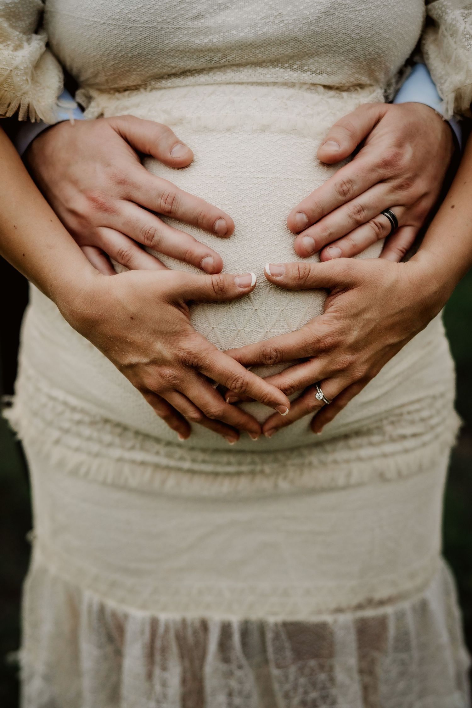 Close-up of hands forming heart shape on pregnant belly in white lace dress.