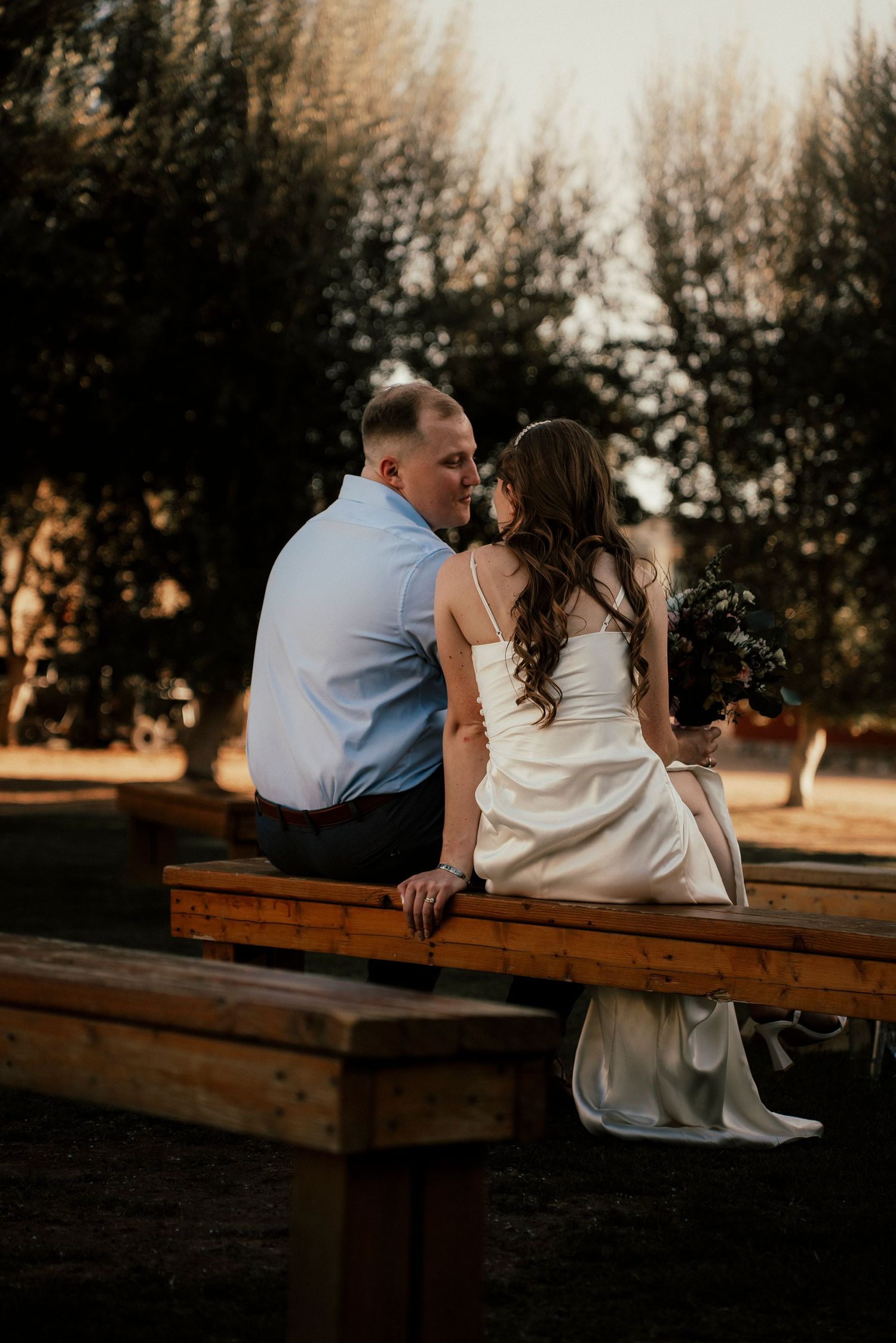 A couple sits together on a rustic wooden bench during sunset for their wedding photoshoot.
