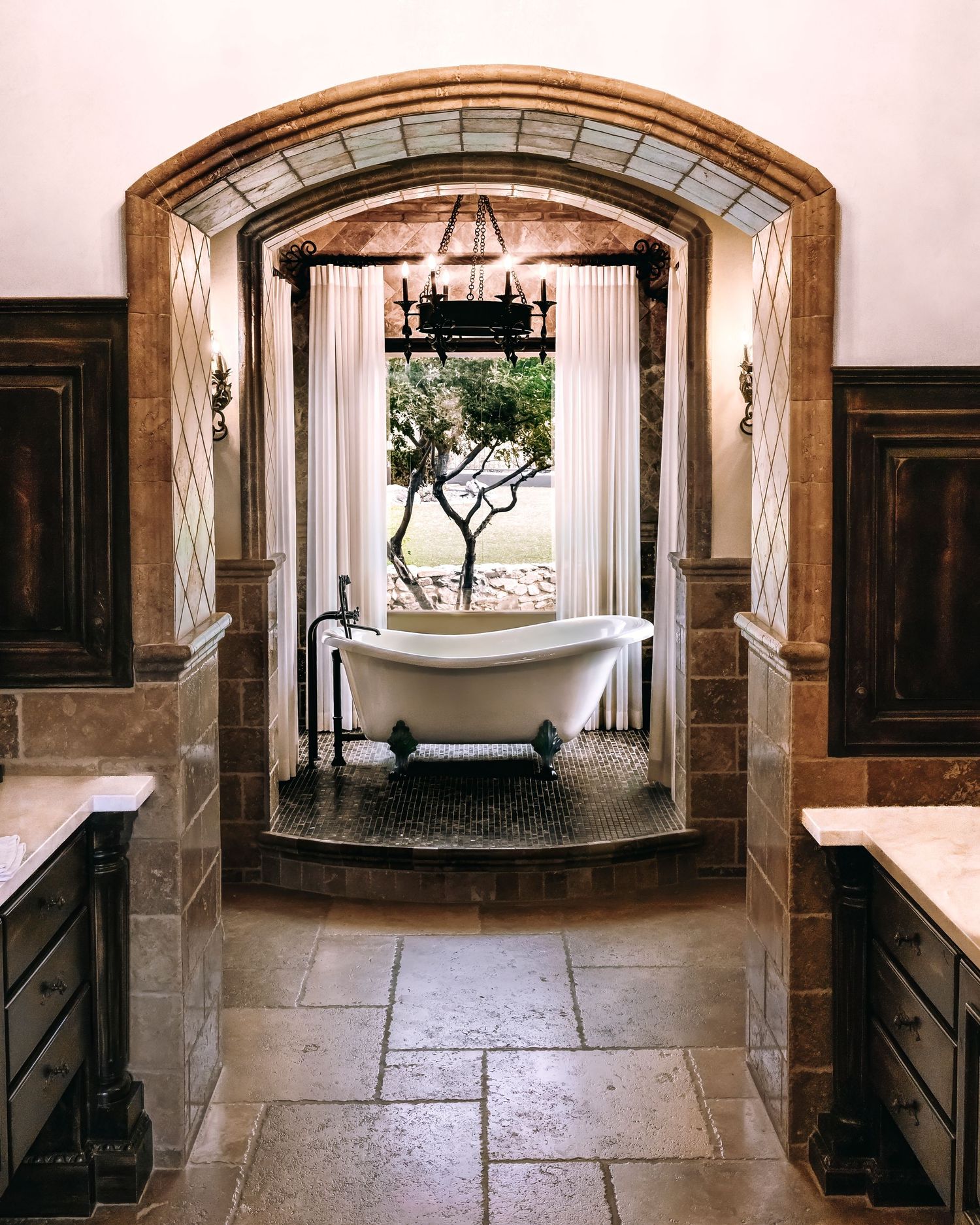 Elegant freestanding clawfoot bathtub framed by arched doorway with vintage chandelier and wood paneled walls.