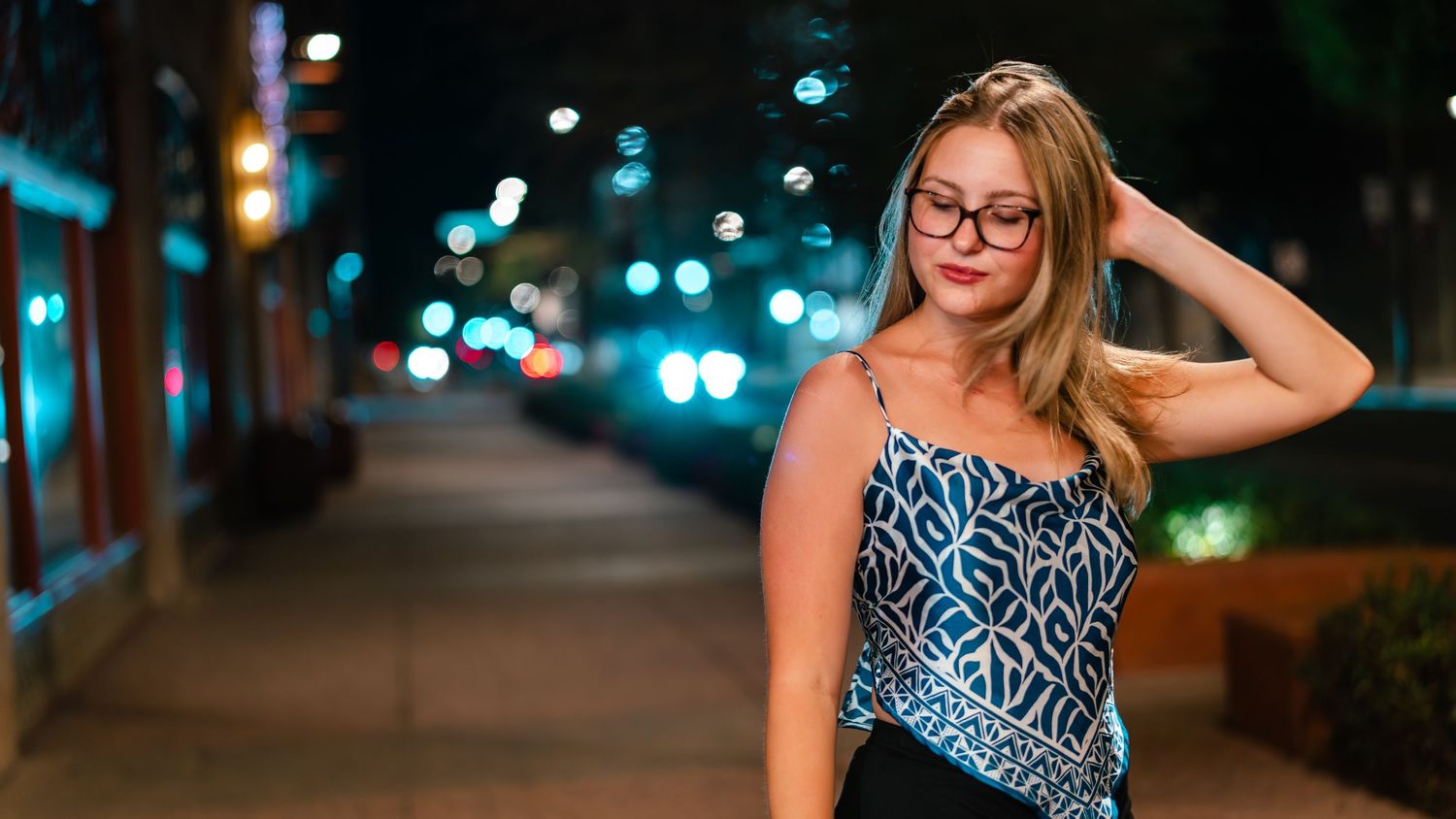 A woman in a blue and white patterned dress poses in an outdoor nighttime city setting.