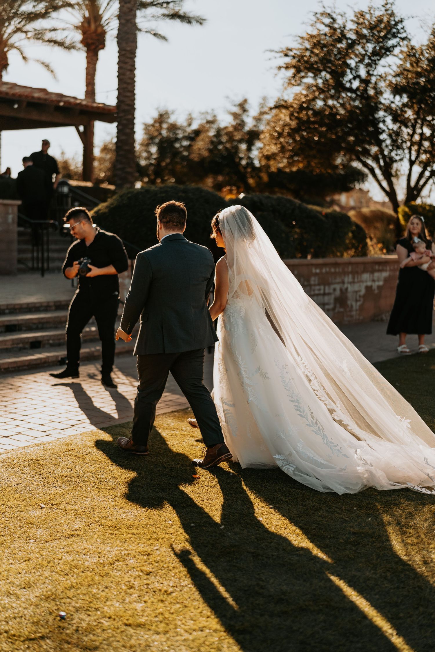 Wedding party walks along outdoor path during golden sunset with long bridal veil trailing behind.