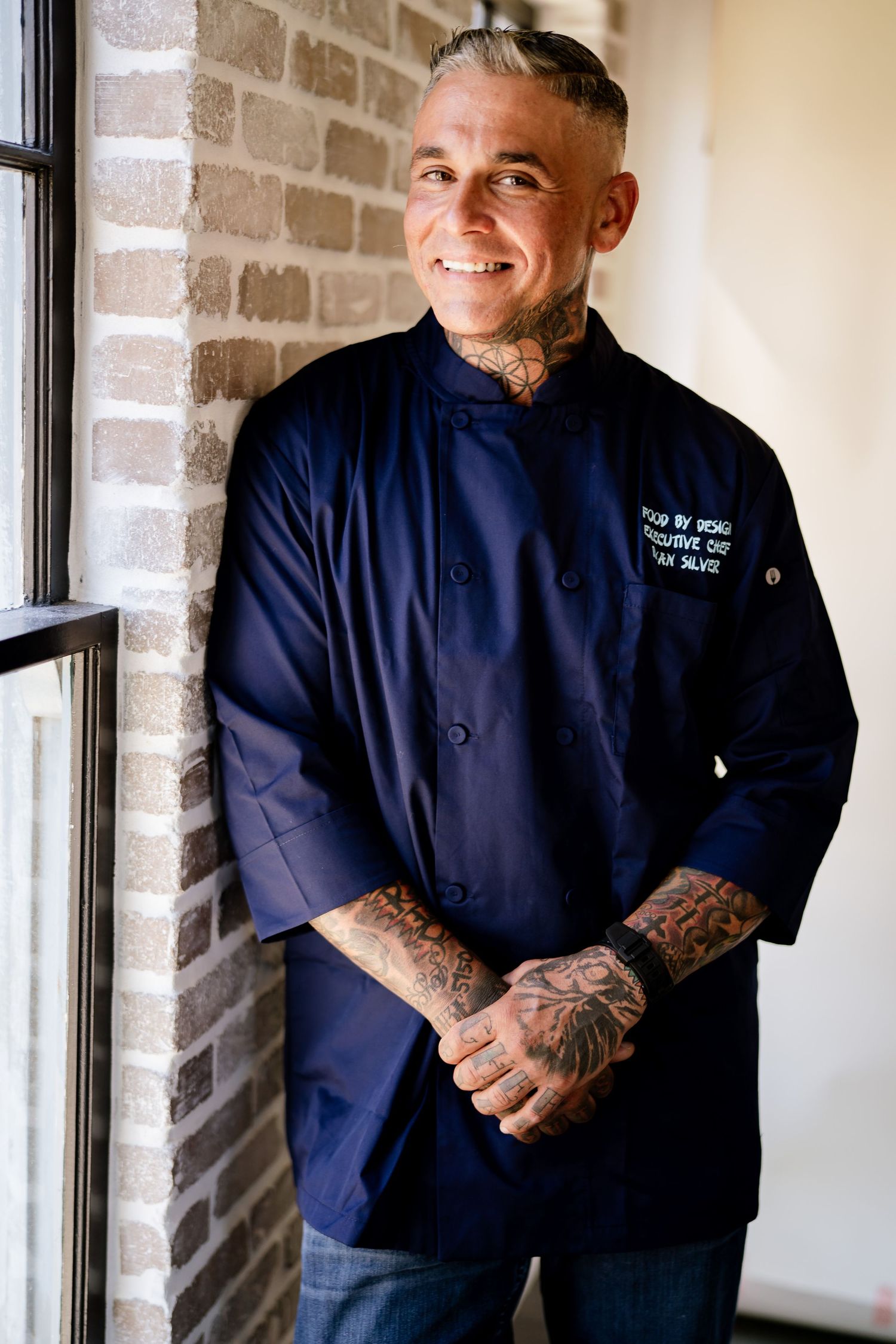 Chef in navy blue uniform with tattoos stands by brick wall.