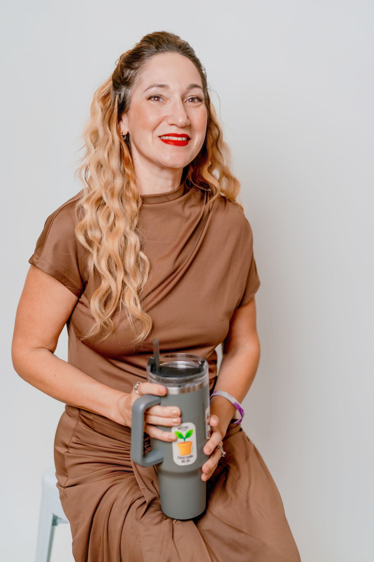 Woman in brown dress laughs while holding large gray tumbler against light background.