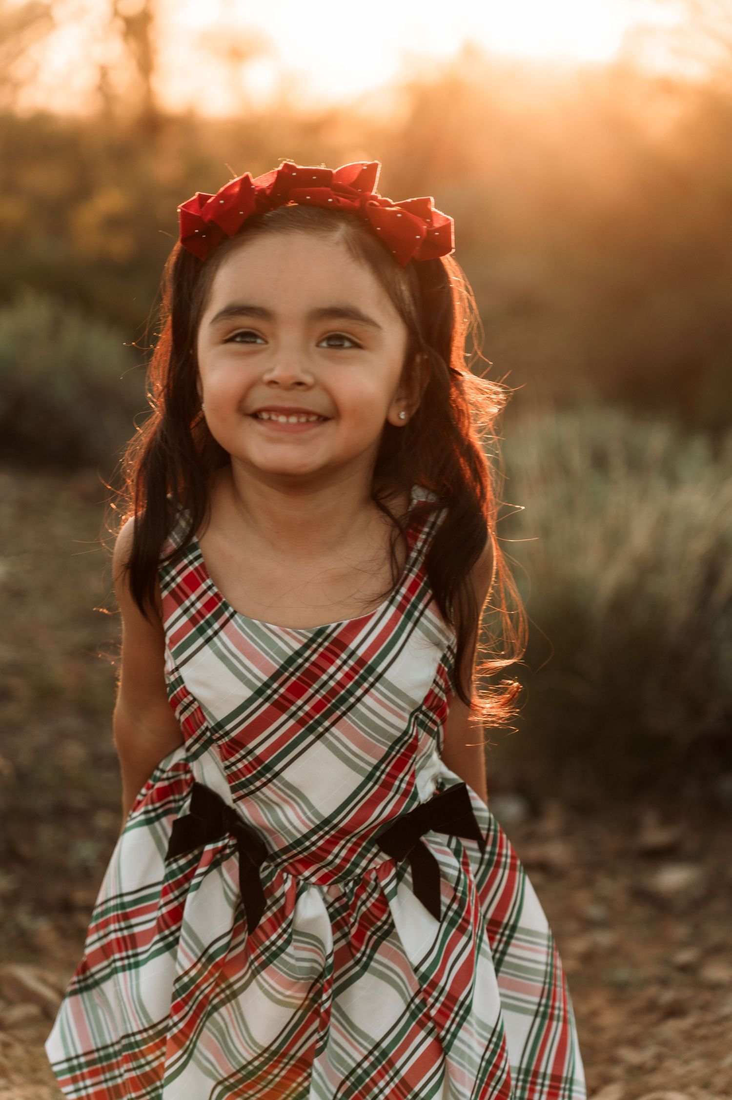 A young child wearing a red and white plaid dress poses outdoors during golden hour sunset.