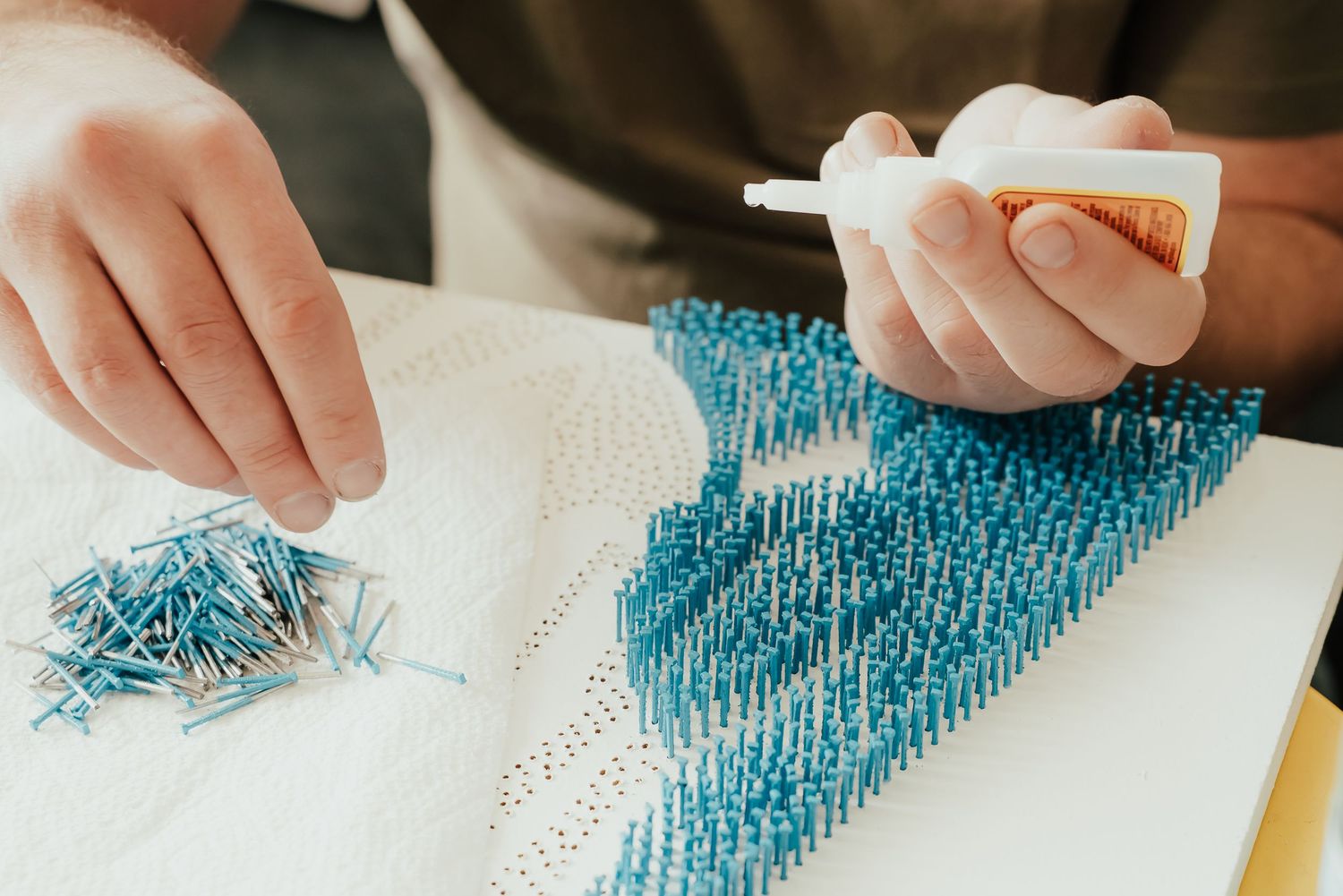 Hands working on a detailed artwork using blue pins on white canvas. Session in Anthem Arizona