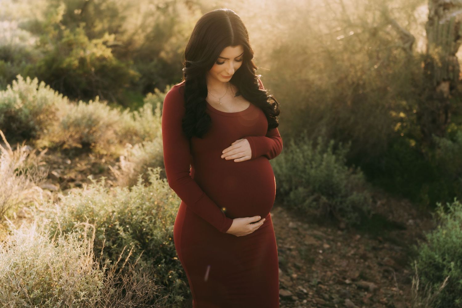 A pregnant woman in a burgundy dress cradles her belly while standing in golden sunlit nature.