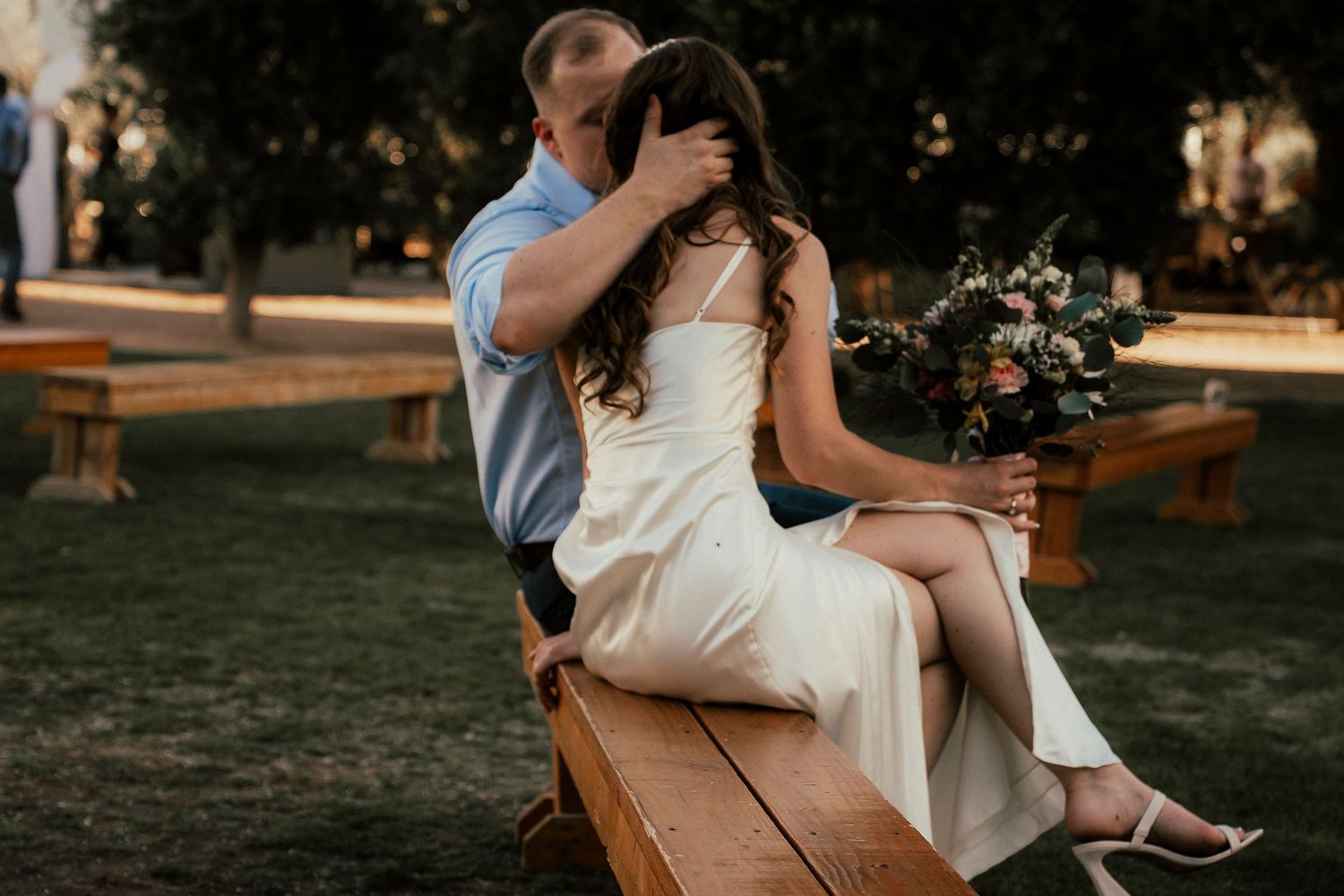 Couple sits intimately on wooden bench during outdoor photoshoot with floral bouquet.