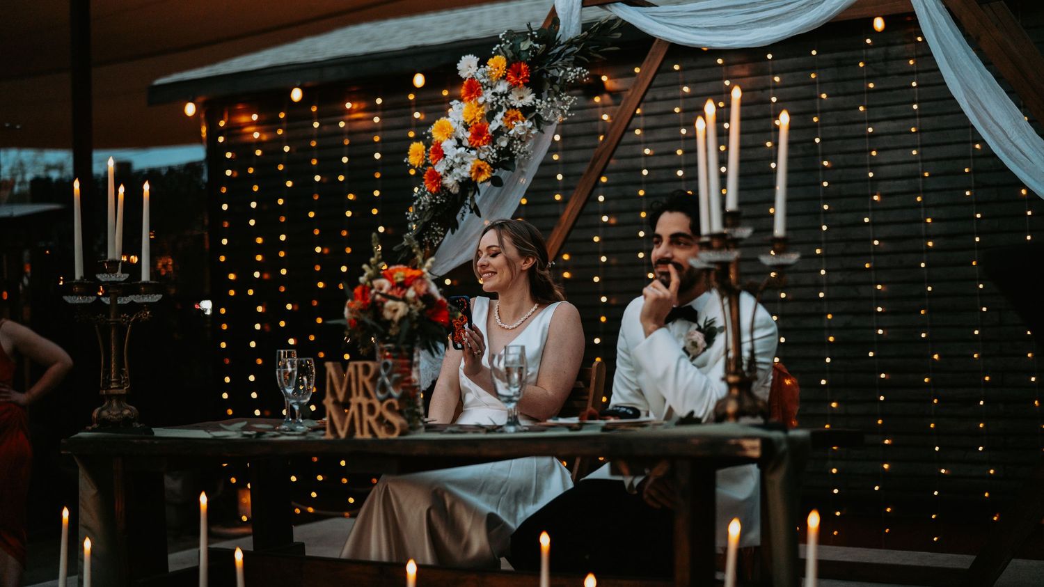 A couple sits at their illuminated reception table decorated with string lights and candles.