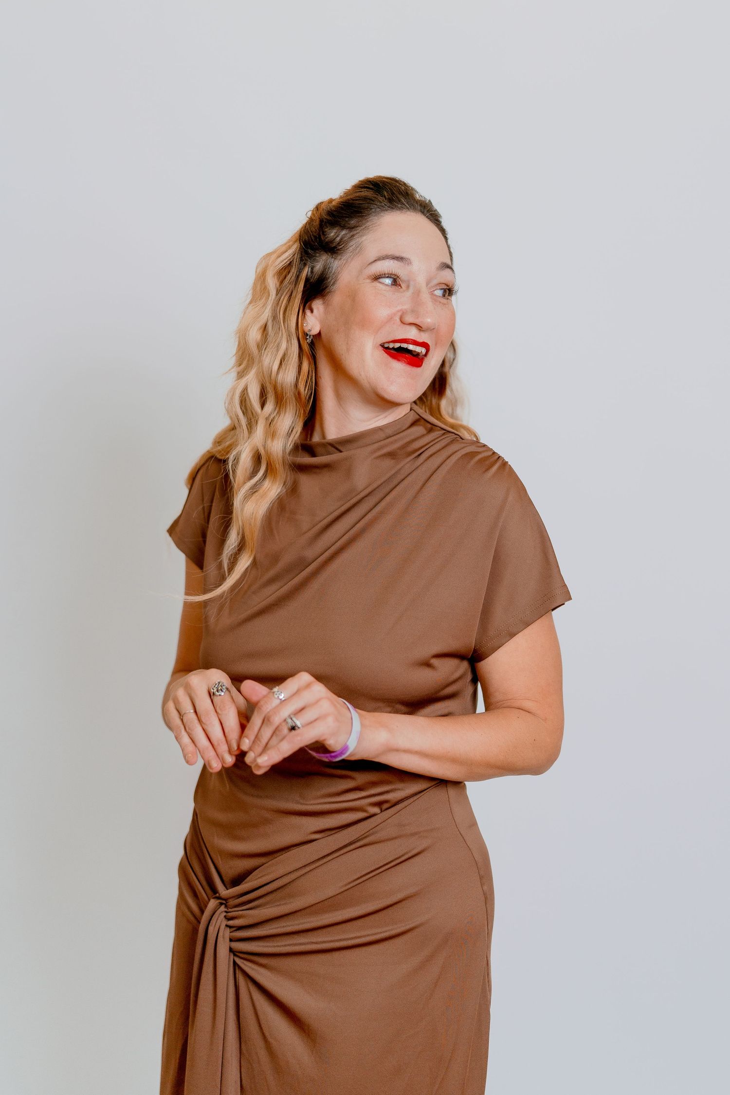 Woman in brown ruched dress smiles joyfully against neutral gray backdrop.