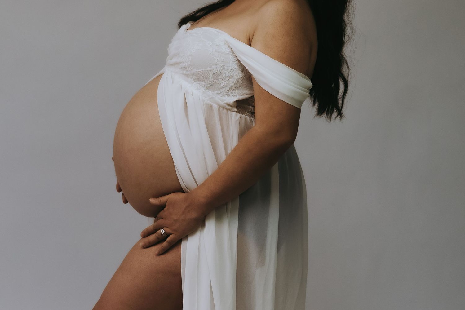 A serene maternity photoshoot showing a pregnant woman in white dress against grey background.