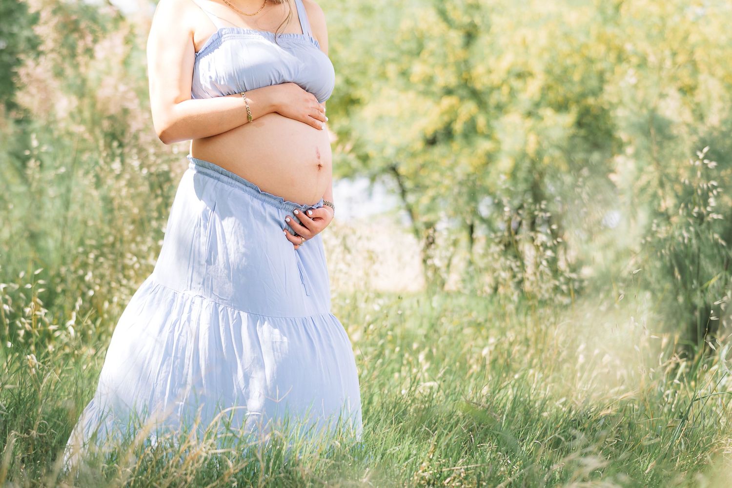 Close-up shot of a pregnant belly in a light blue dress against a dreamy outdoor backdrop.