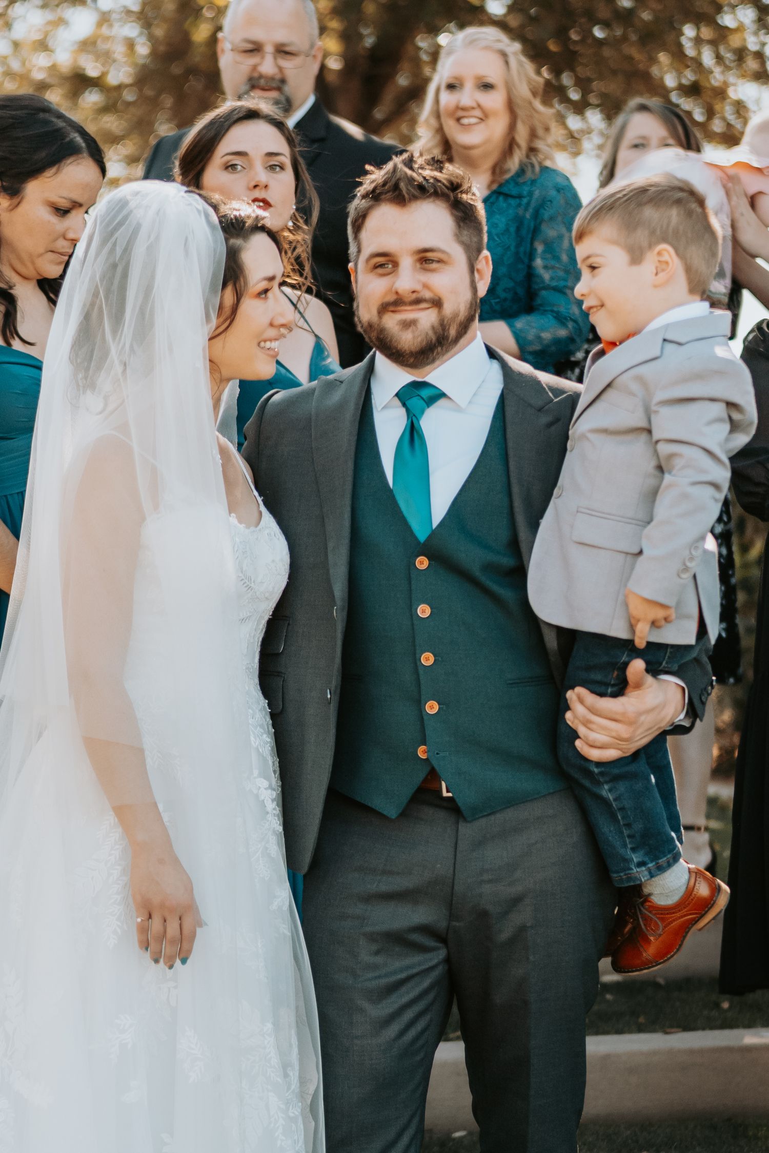 Wedding party in charcoal suits and teal accents poses together with bride in white gown.