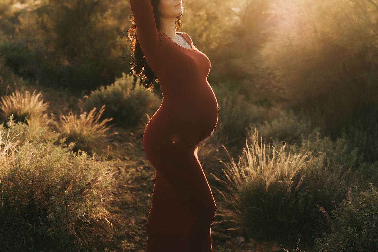 Silhouette of a person in a flowing dress stands against golden sunlit grass during a dreamy sunset photoshoot.