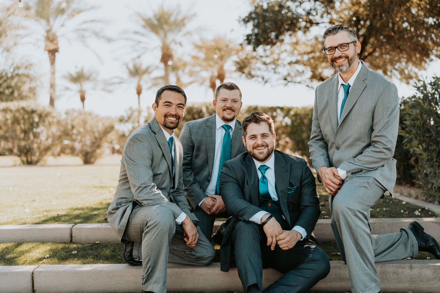 Groomsmen in gray suits and teal ties share candid laughs while sitting together outdoors.