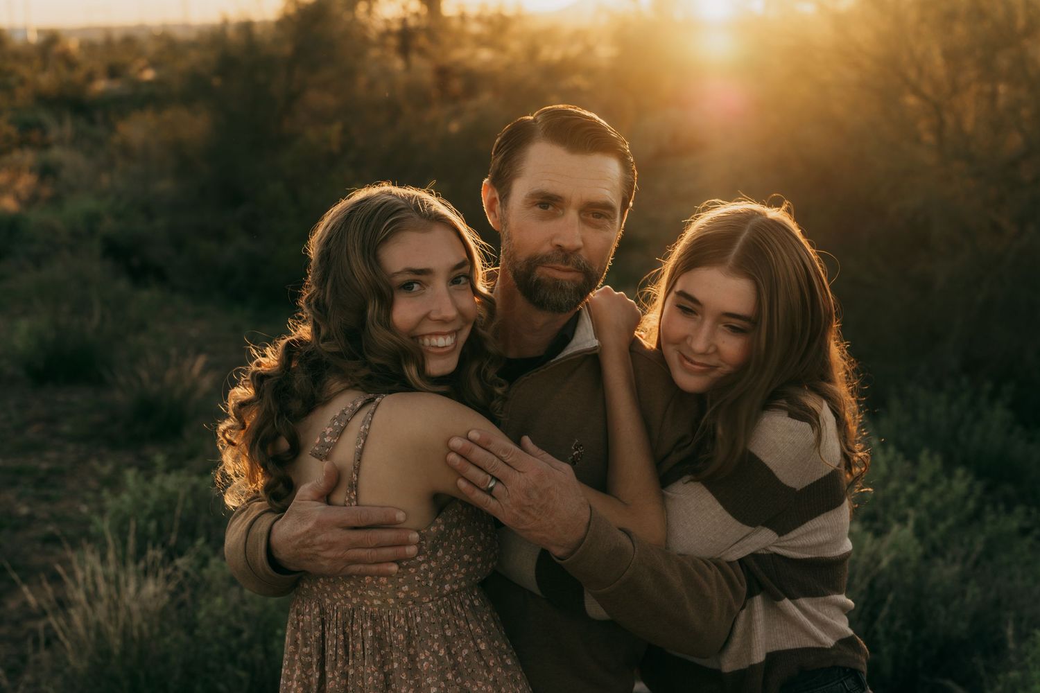 Three people embrace and smile together during golden hour sunset in a natural outdoor setting.