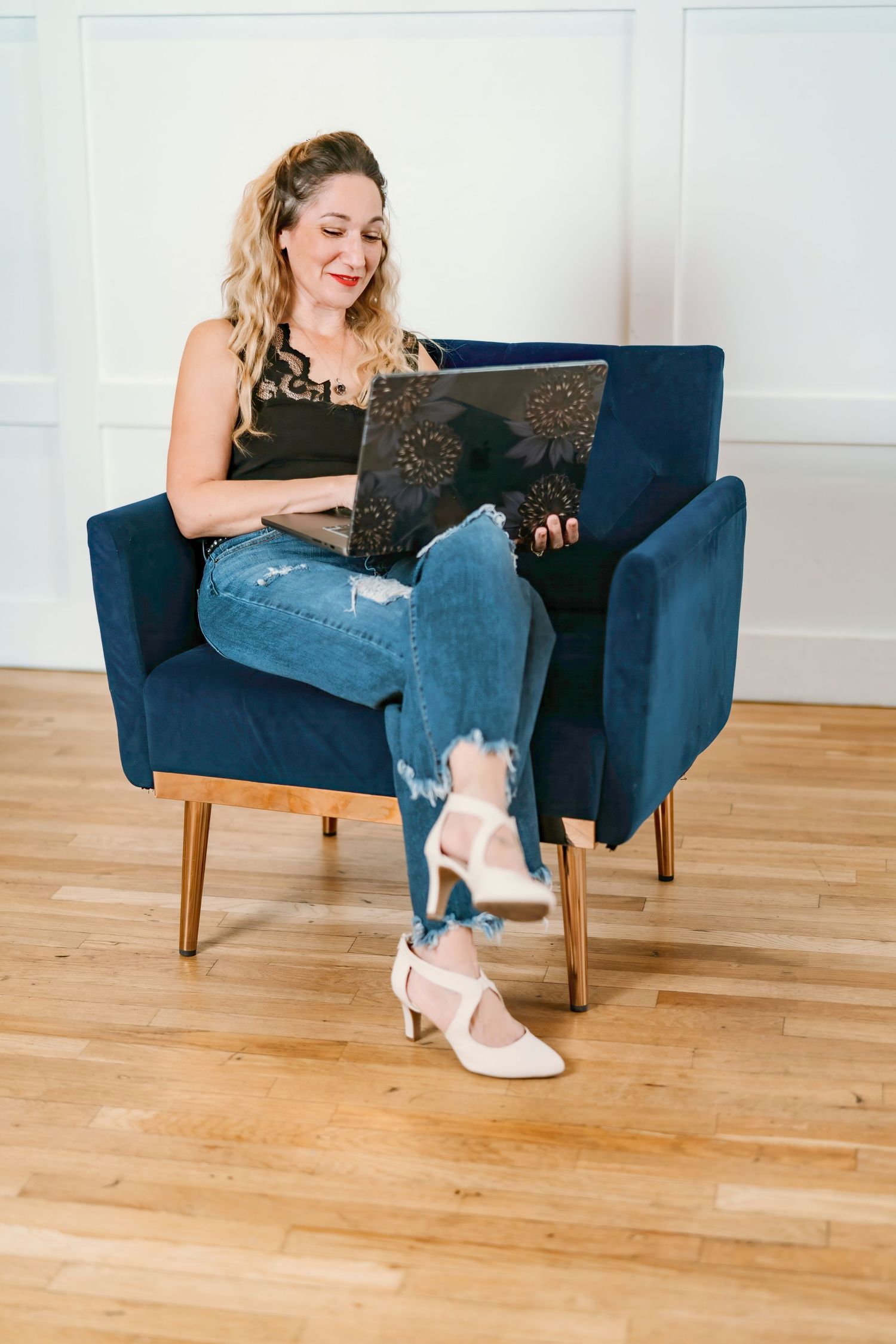 Woman relaxes in a navy blue armchair wearing ripped jeans and pink heels while working on a laptop.