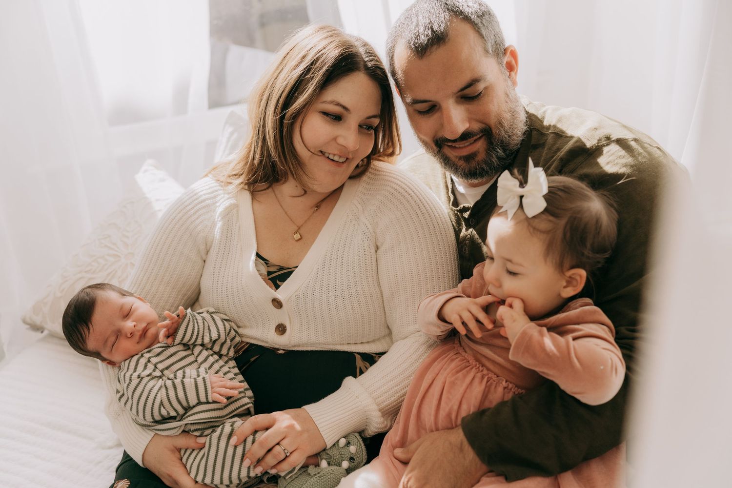 Family cuddles together on white bedding in a bright, naturally lit bedroom during an intimate lifestyle photo session.