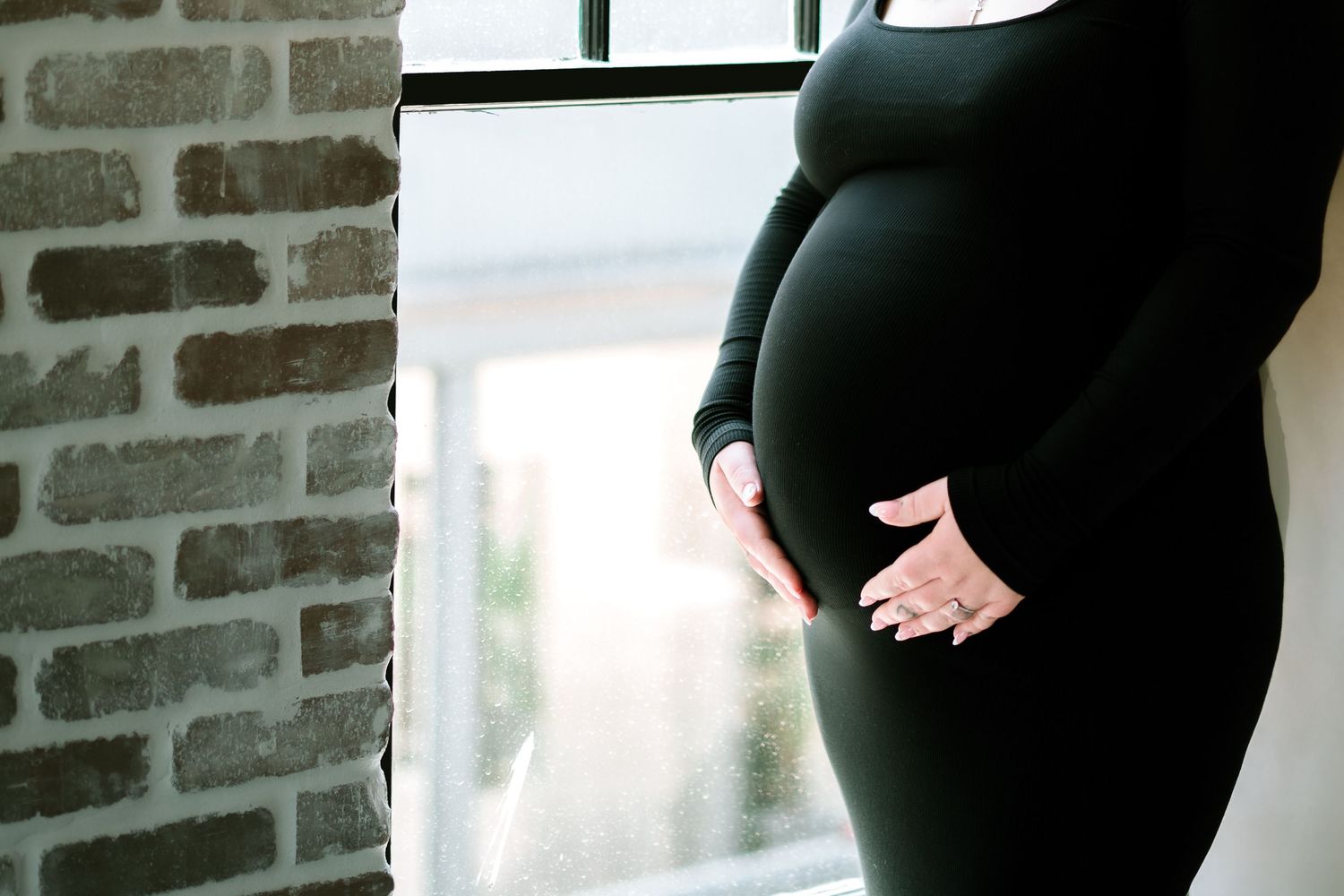A silhouette of a pregnant woman in a black dress stands near a brick wall by a window.