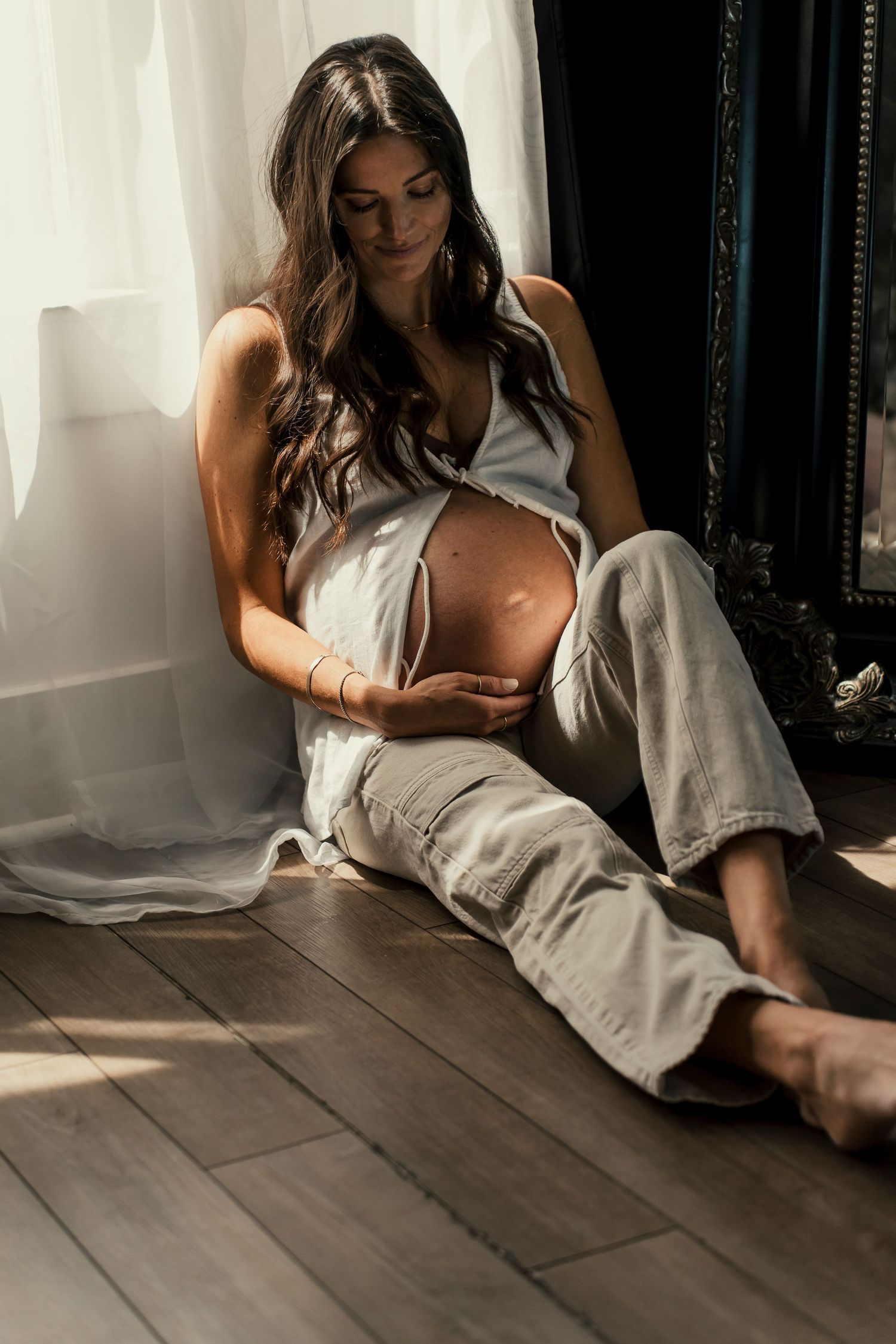 Woman in light dress lounging on wooden floor in sunlit room with white curtains.