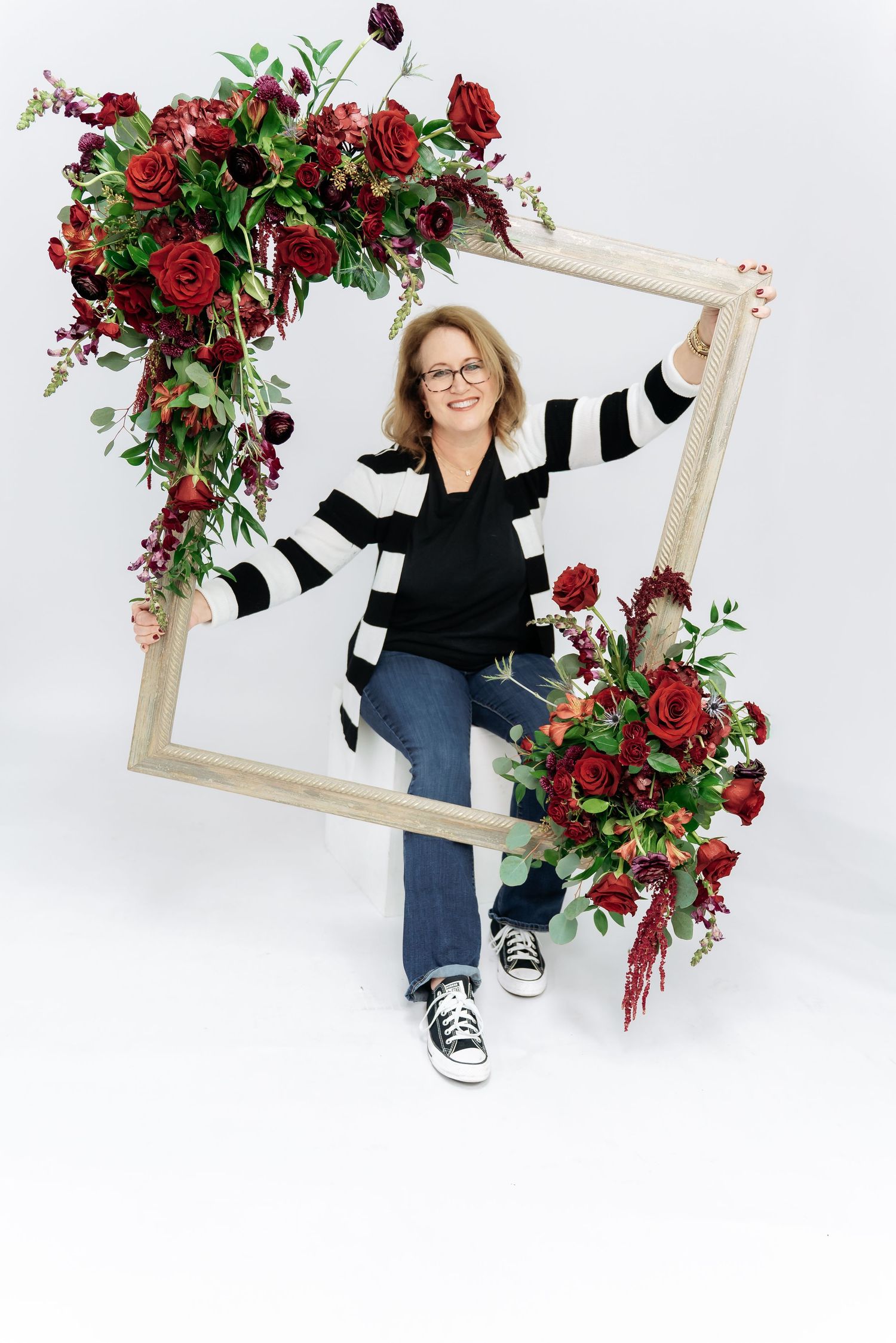 Decorative wooden frame adorned with lush burgundy roses and greenery arrangements on corners against white background.