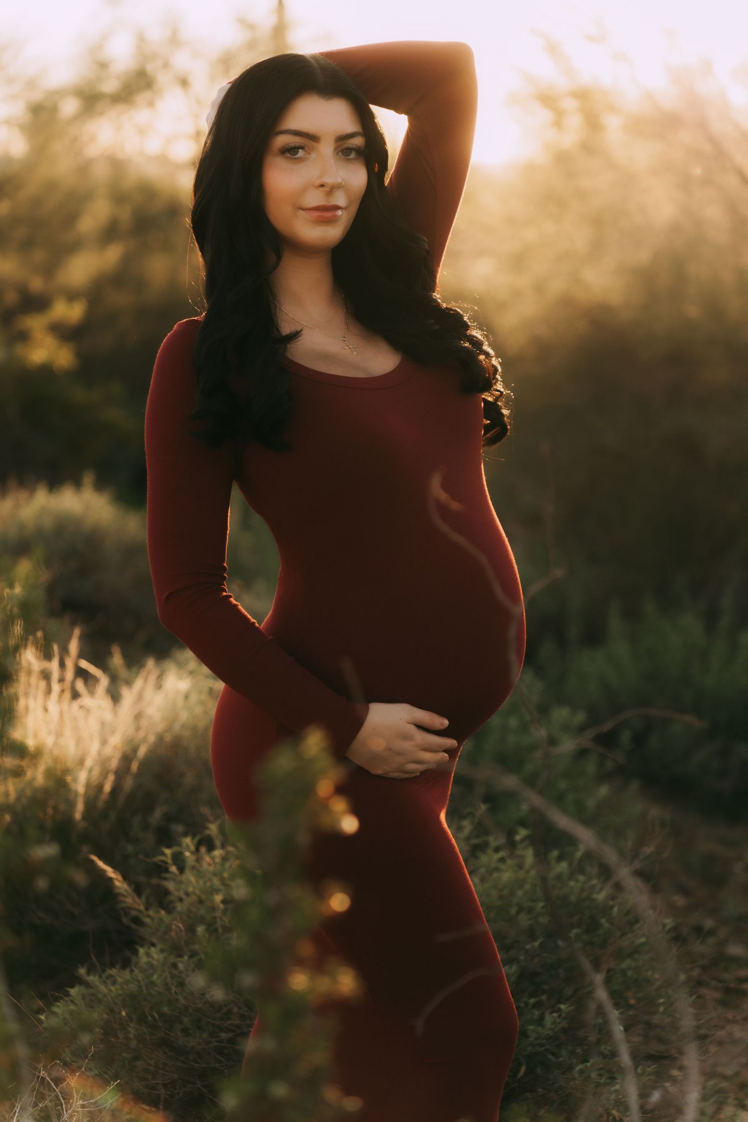 A pregnant woman in a burgundy dress poses in golden sunlight against a natural outdoor backdrop.