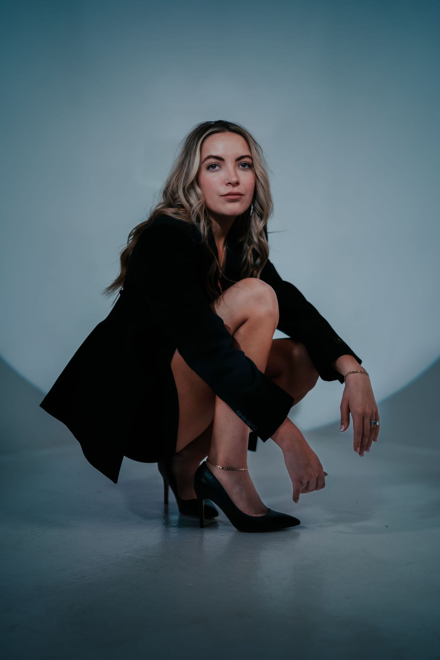 Artistic fashion portrait of model in black blazer and heels crouching on reflective surface with moody lighting.