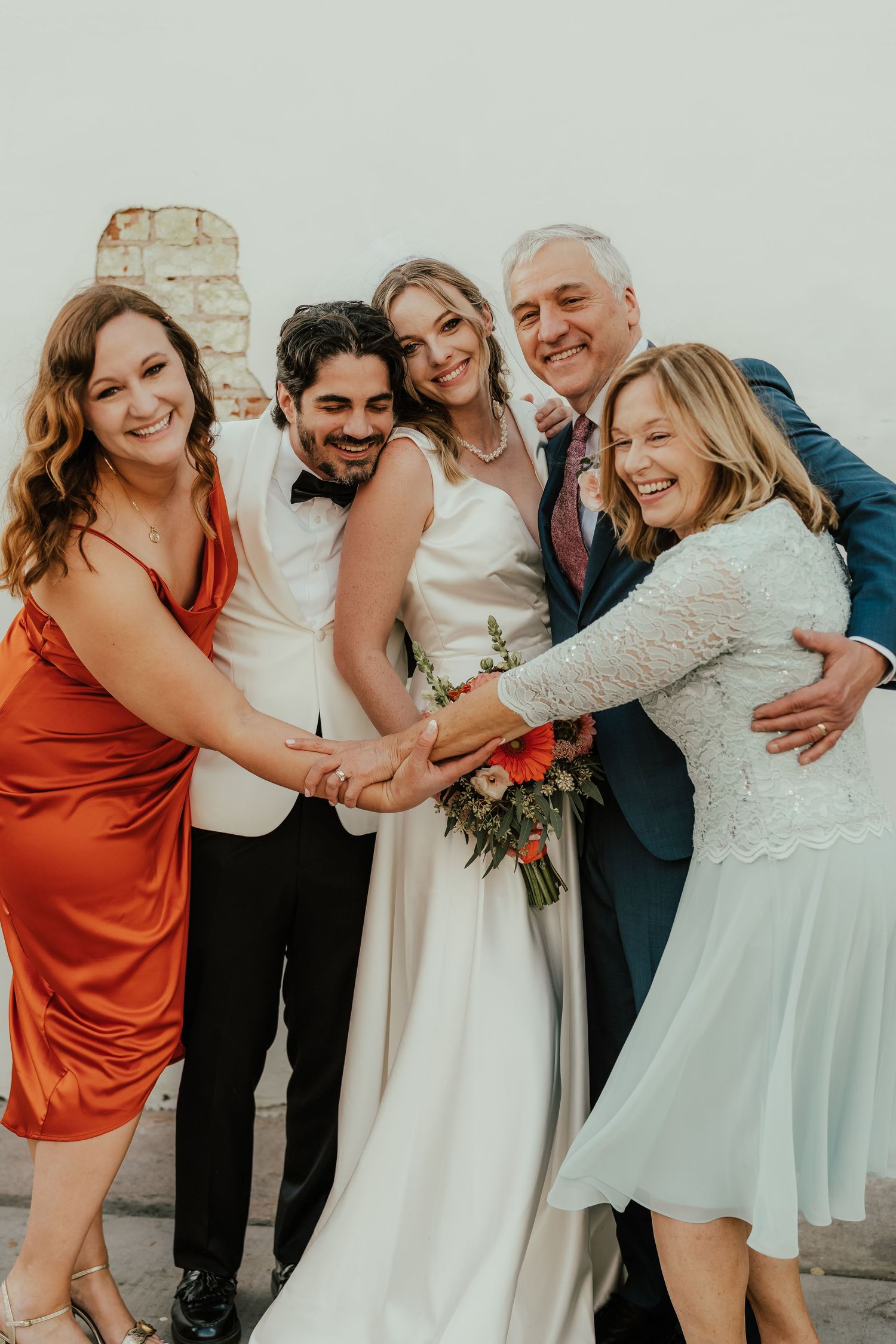 Wedding party in white, teal, and orange attire hugs and laughs together against light background.