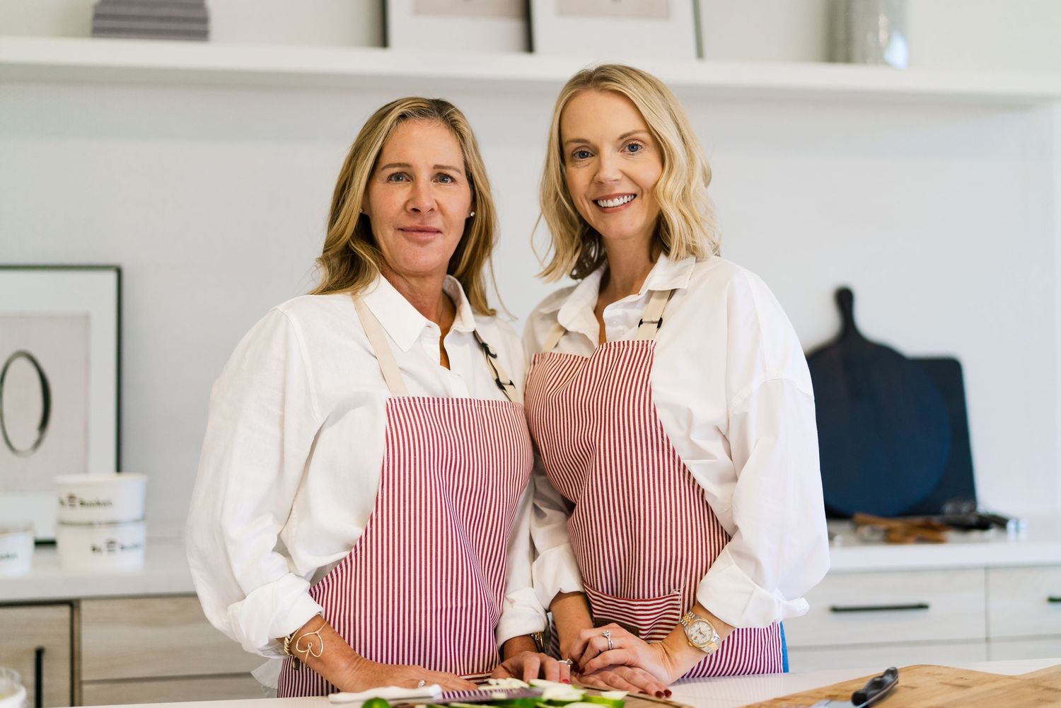 Two women in pink aprons stand together in a bright white kitchen while cooking and preparing food.