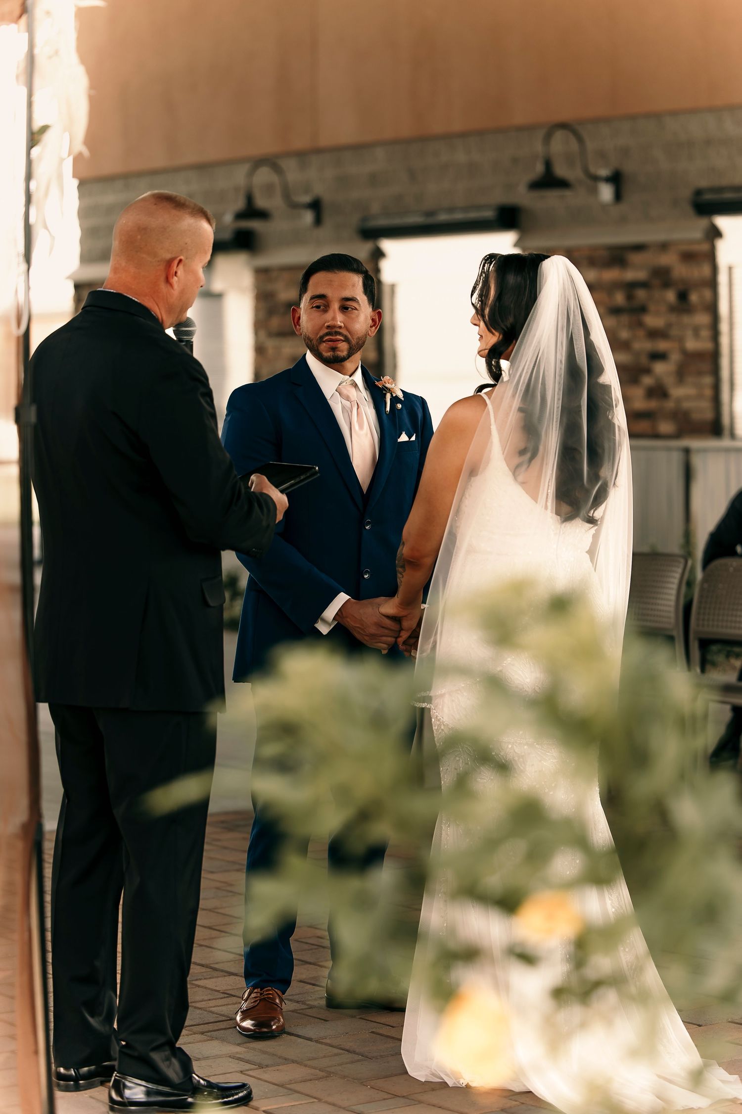 A wedding ceremony takes place on an outdoor patio with brick pillars in the background.
