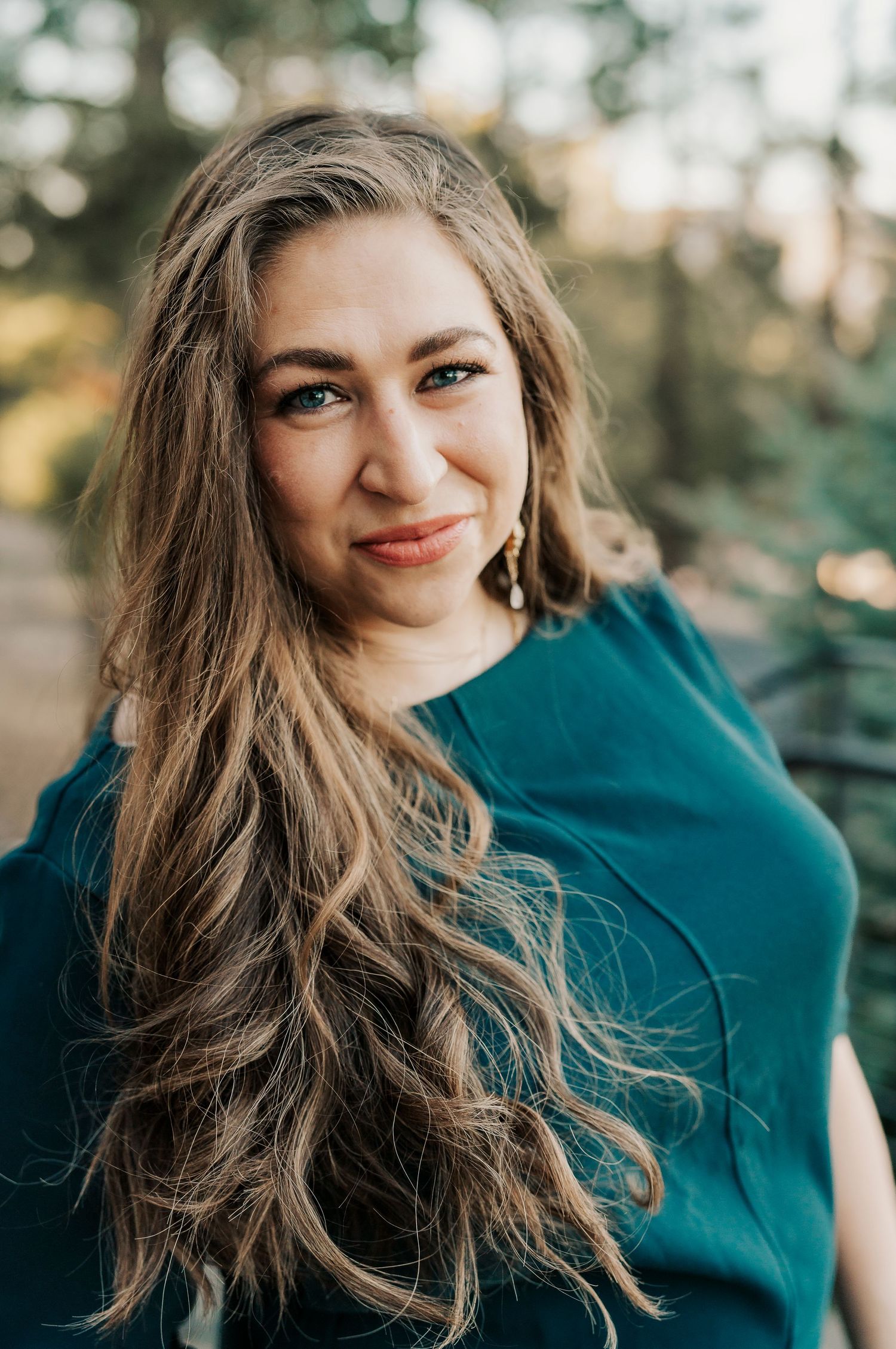 Portrait of a person in a teal top with flowing brown hair against a natural outdoor setting.