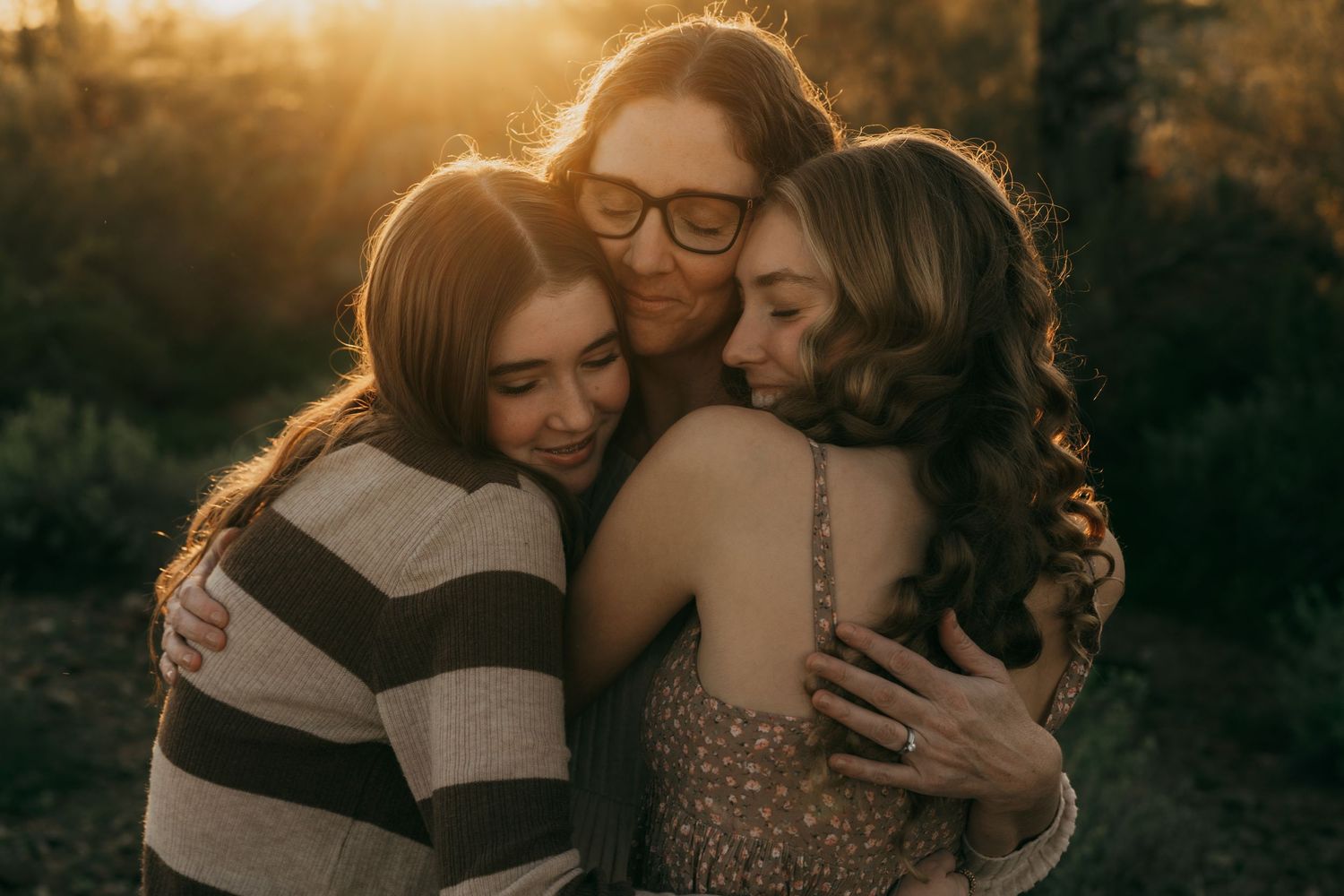 A warm sunset moment captures three people hugging closely in an outdoor setting with golden lighting.