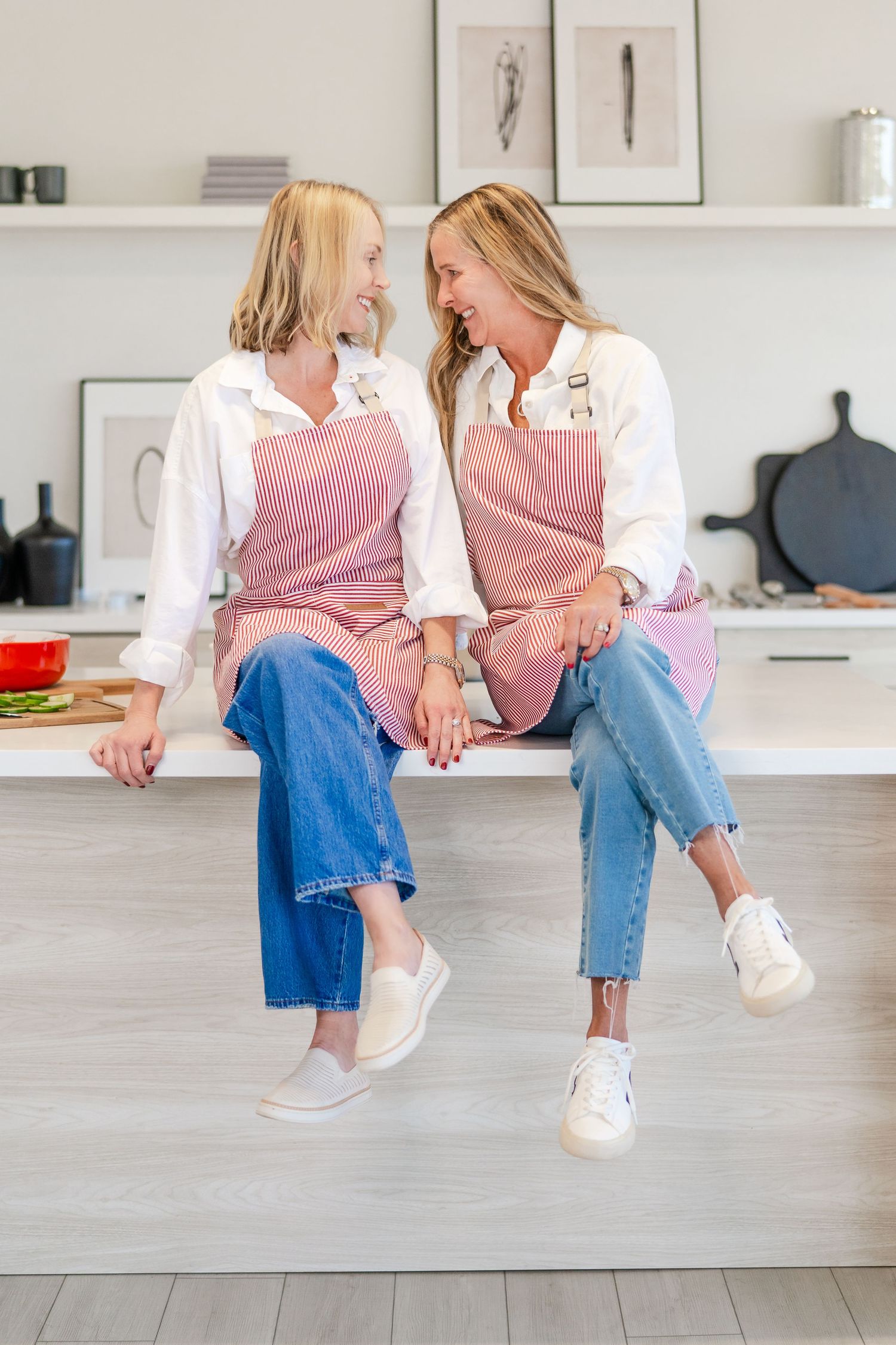 Two people in matching pink shirts and jeans sit casually on a white counter in a modern minimalist kitchen setting.