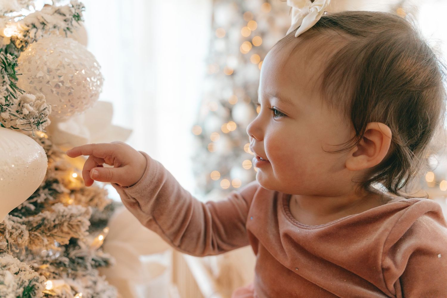 A baby in a pink sweater reaches for glittering holiday decorations in a soft, warm-lit setting.