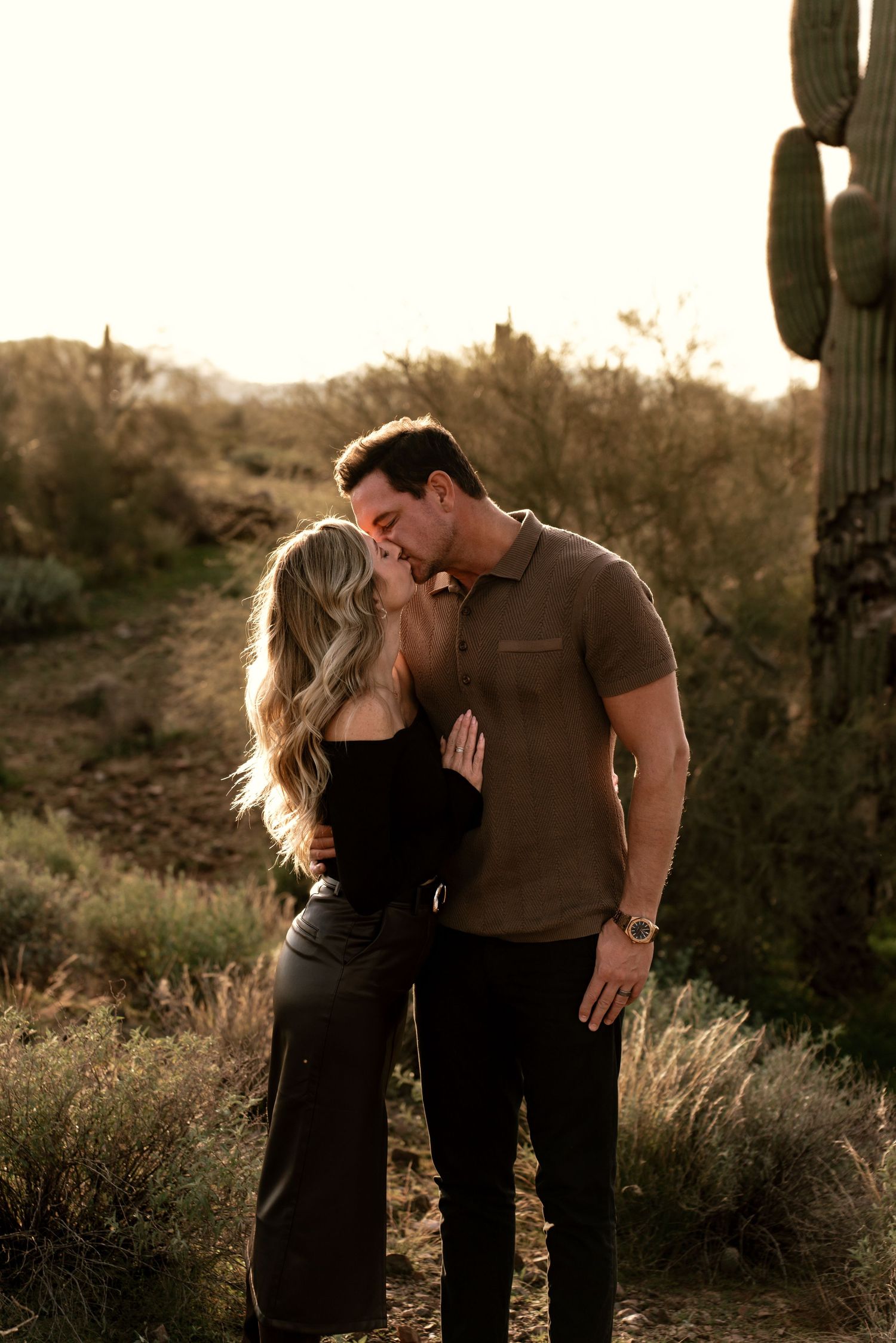 A romantic couple embraces at sunset in a desert landscape with saguaro cacti and golden light.