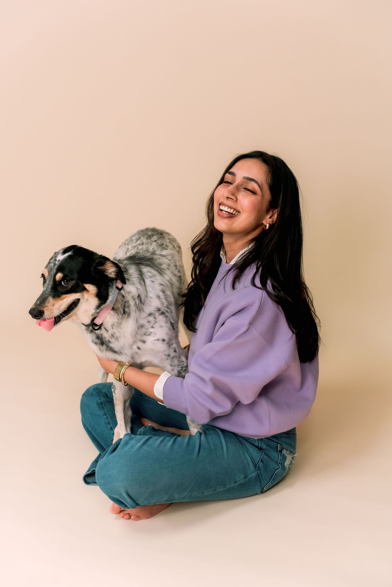 Person in purple sweater and teal pants sits cross-legged with a happy spotted dog against beige background.