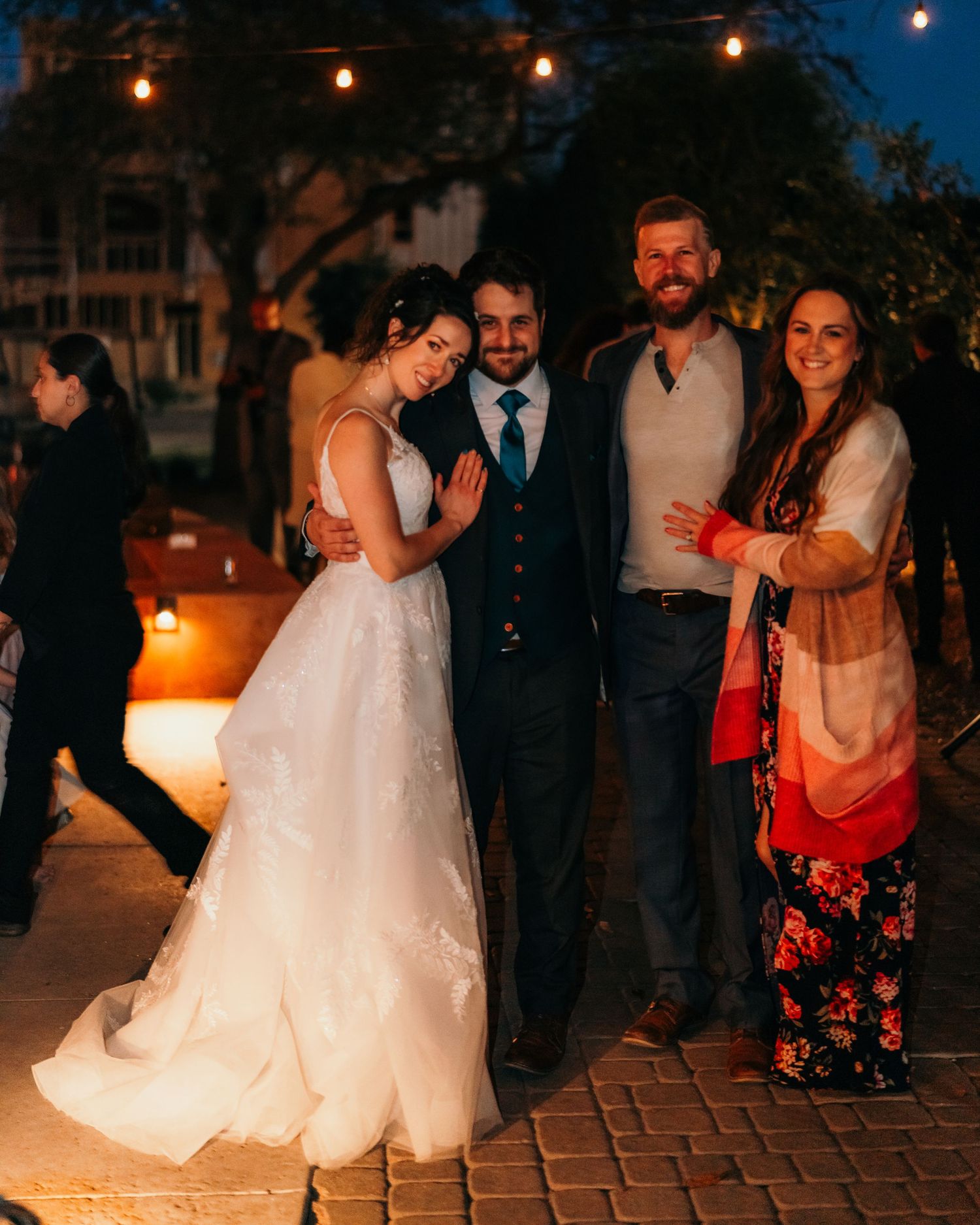 Wedding guests gather for evening photos on a brick patio under string lights.