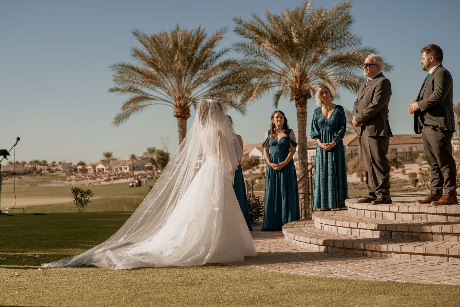 Wedding ceremony takes place outdoors on a sunny day with palm trees and a golf course backdrop.