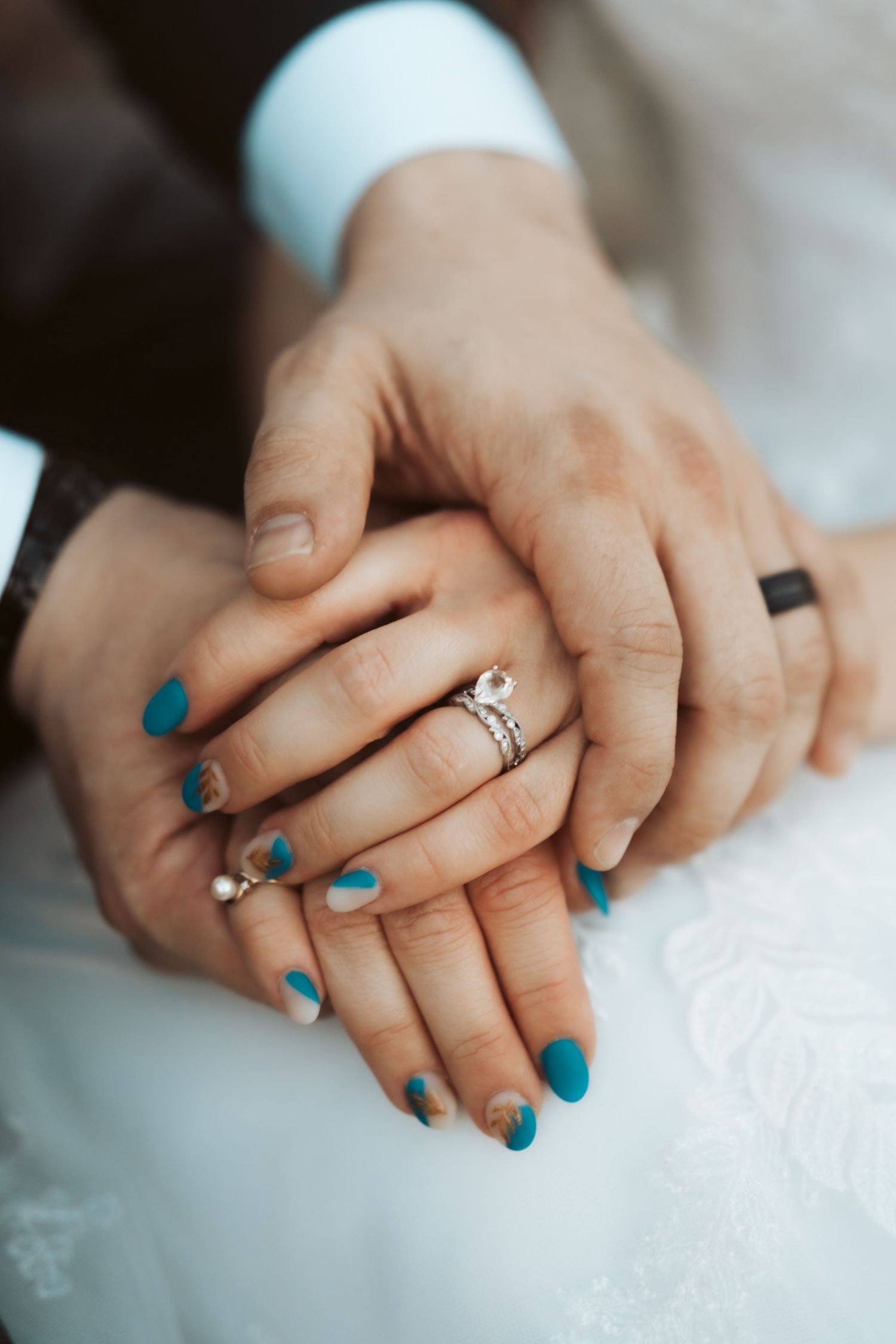 Close-up of hands showing engagement ring and turquoise nail polish against white dress fabric.