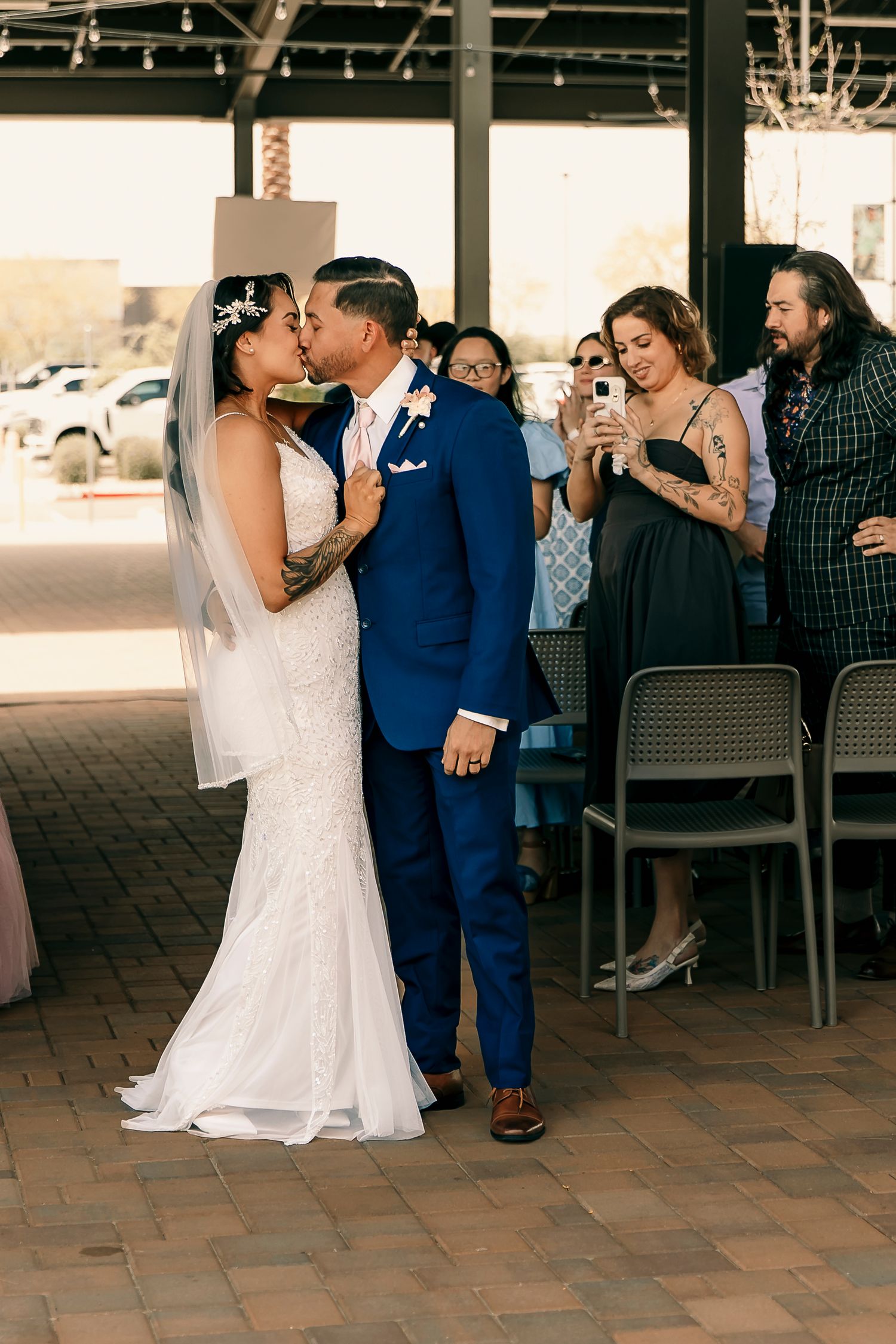 A newlywed couple shares a kiss while guests capture the moment on their phones.