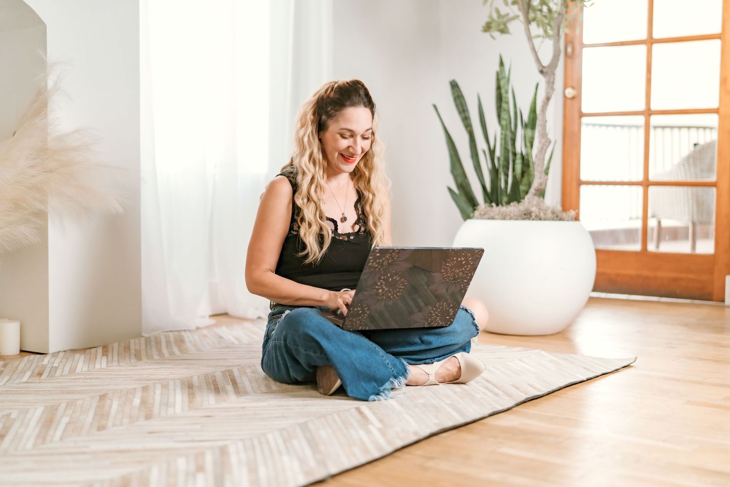 Person sits cross-legged on floor using laptop in bright modern room with houseplant and wooden flooring.