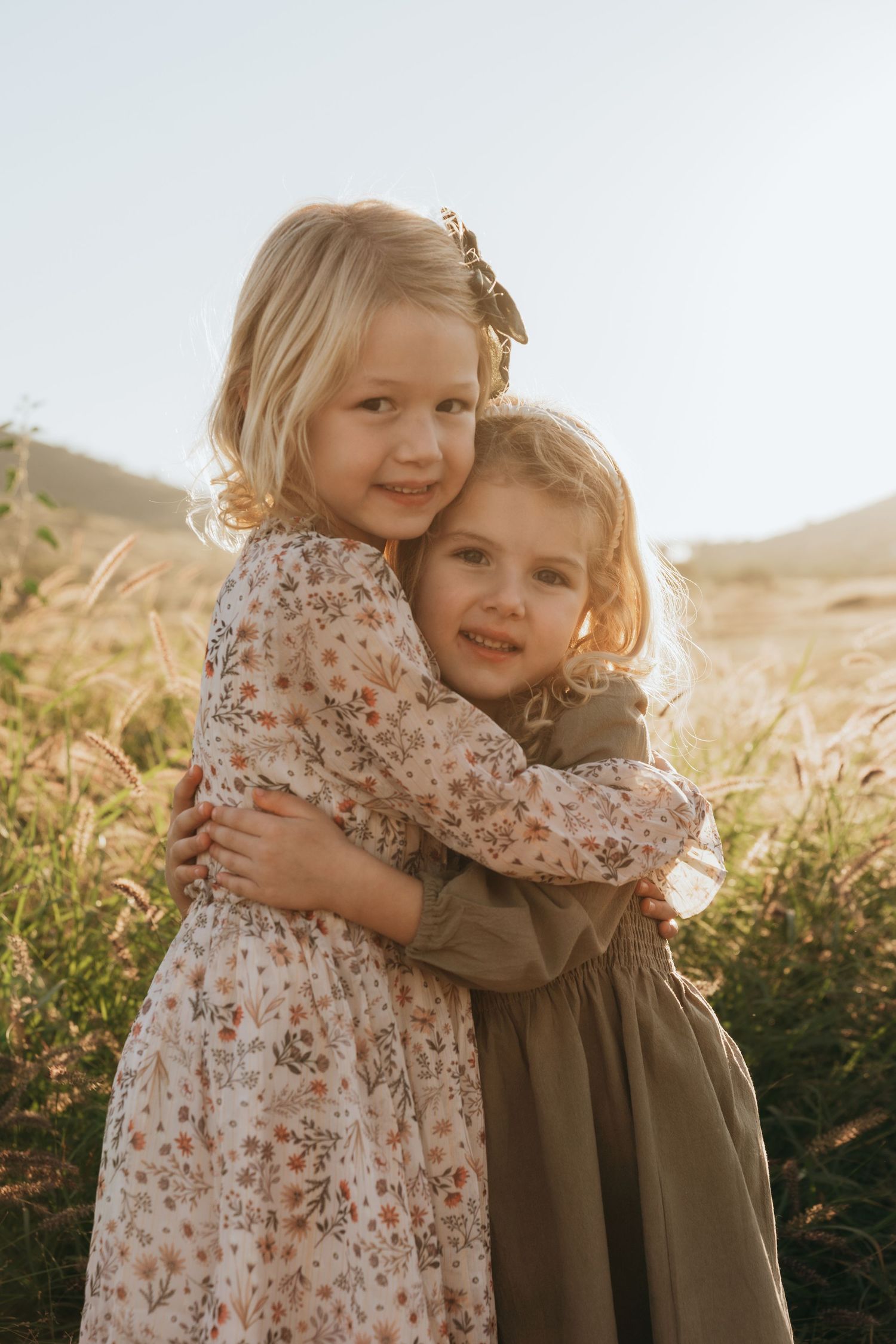 Two young children in floral and olive dresses embrace while smiling in a sunlit meadow at golden hour.