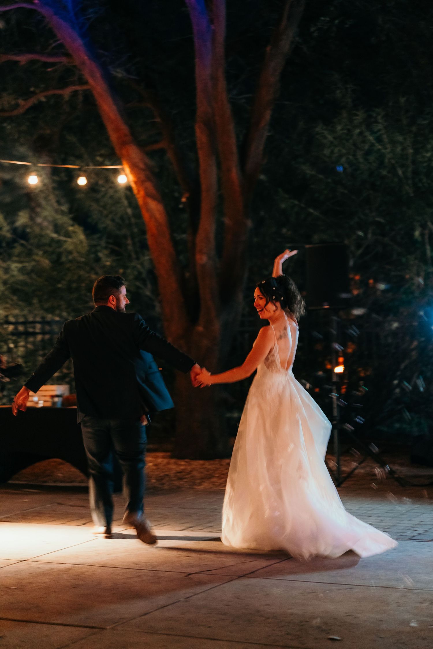 A couple shares their first dance on an outdoor patio illuminated by string lights.