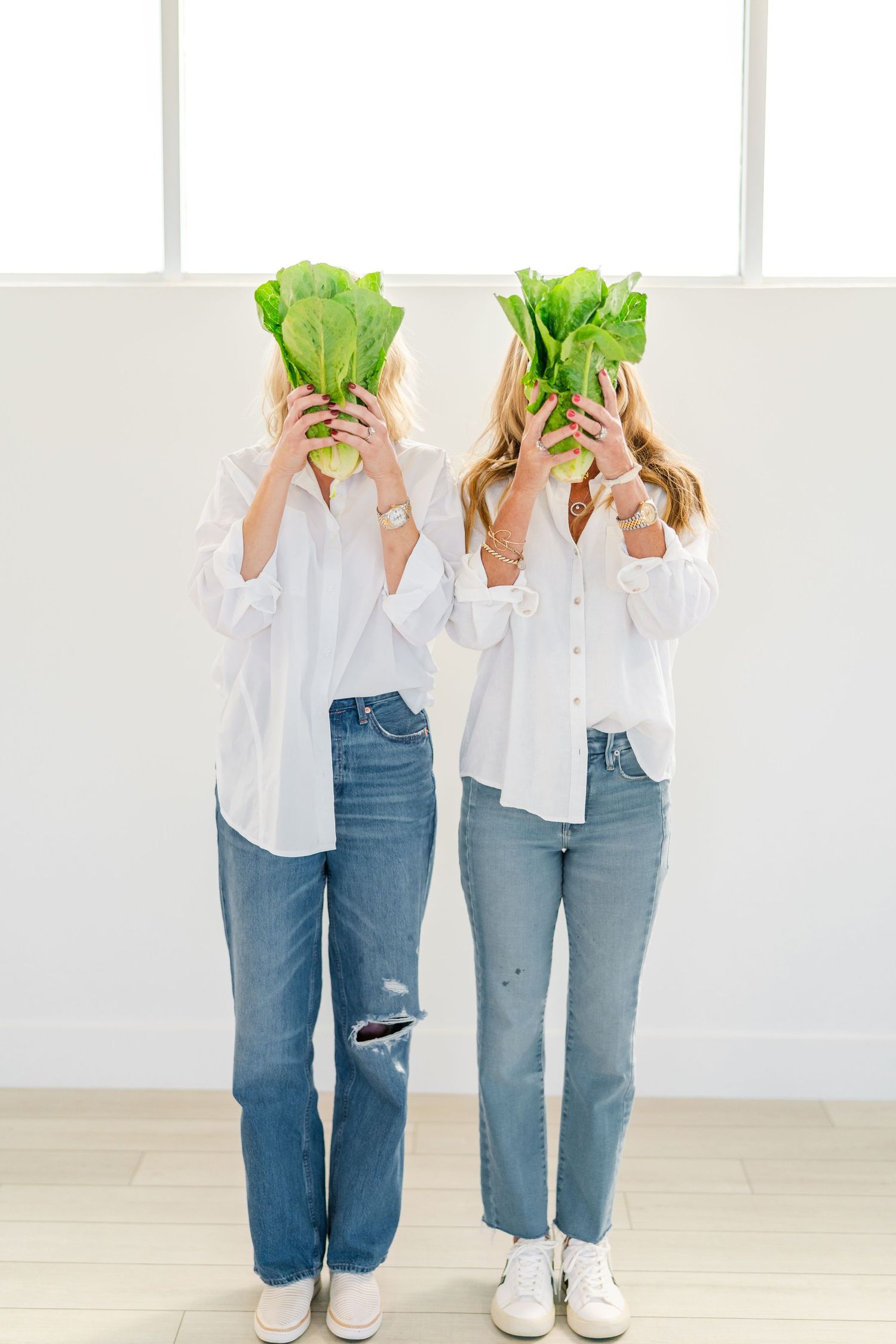 Two people in white shirts and jeans hold fresh lettuce heads in front of their faces while standing against a white wall.