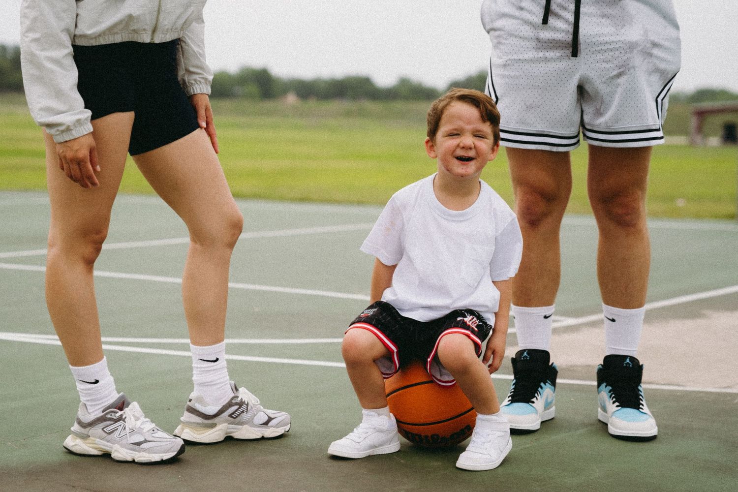 A laughing child sits on a basketball on an outdoor court, flanked by two adults in athletic wear and sneakers.