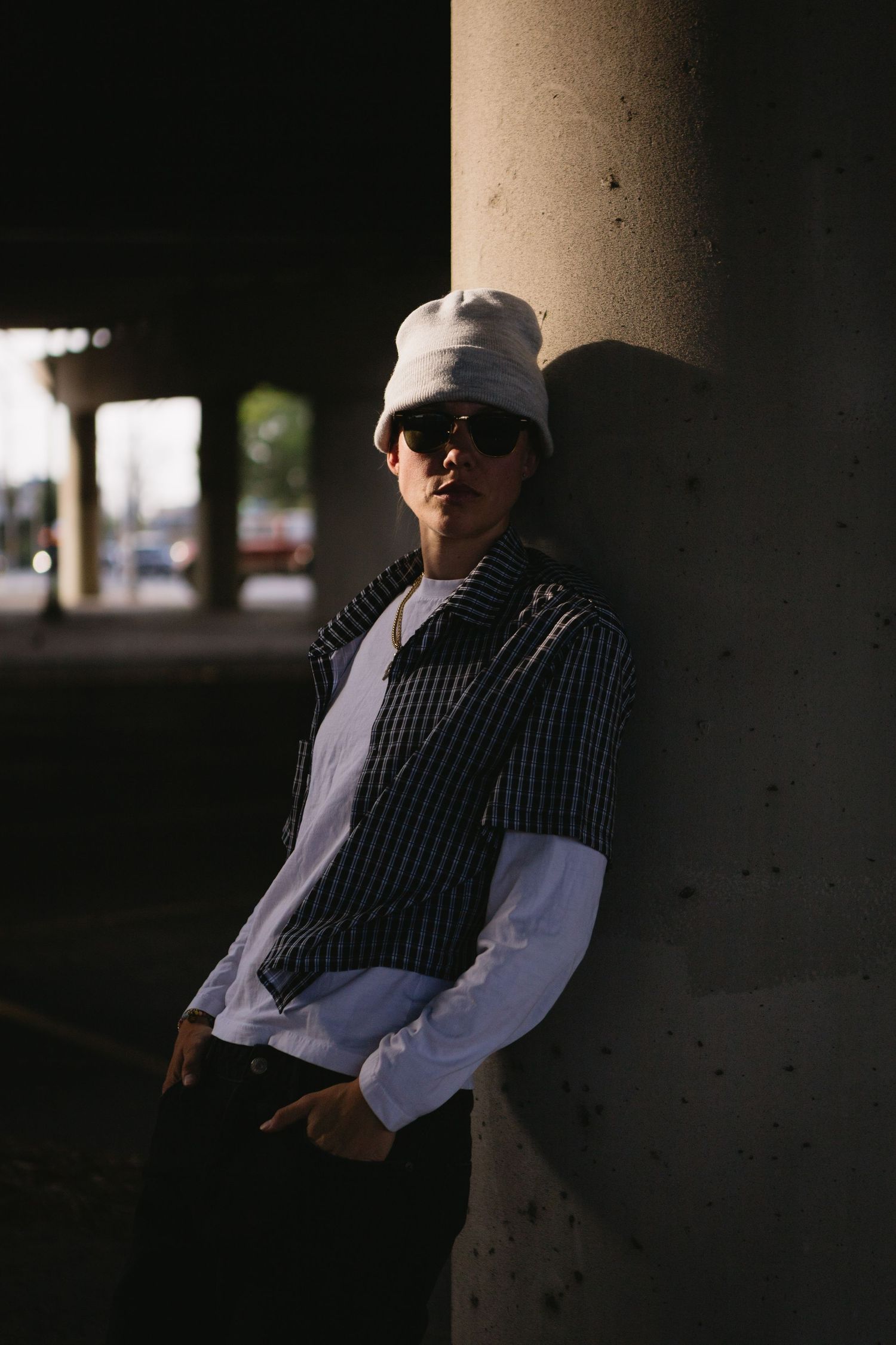 Young person in white bucket hat and sunglasses leaning against concrete pillar under bridge in striped vest.
