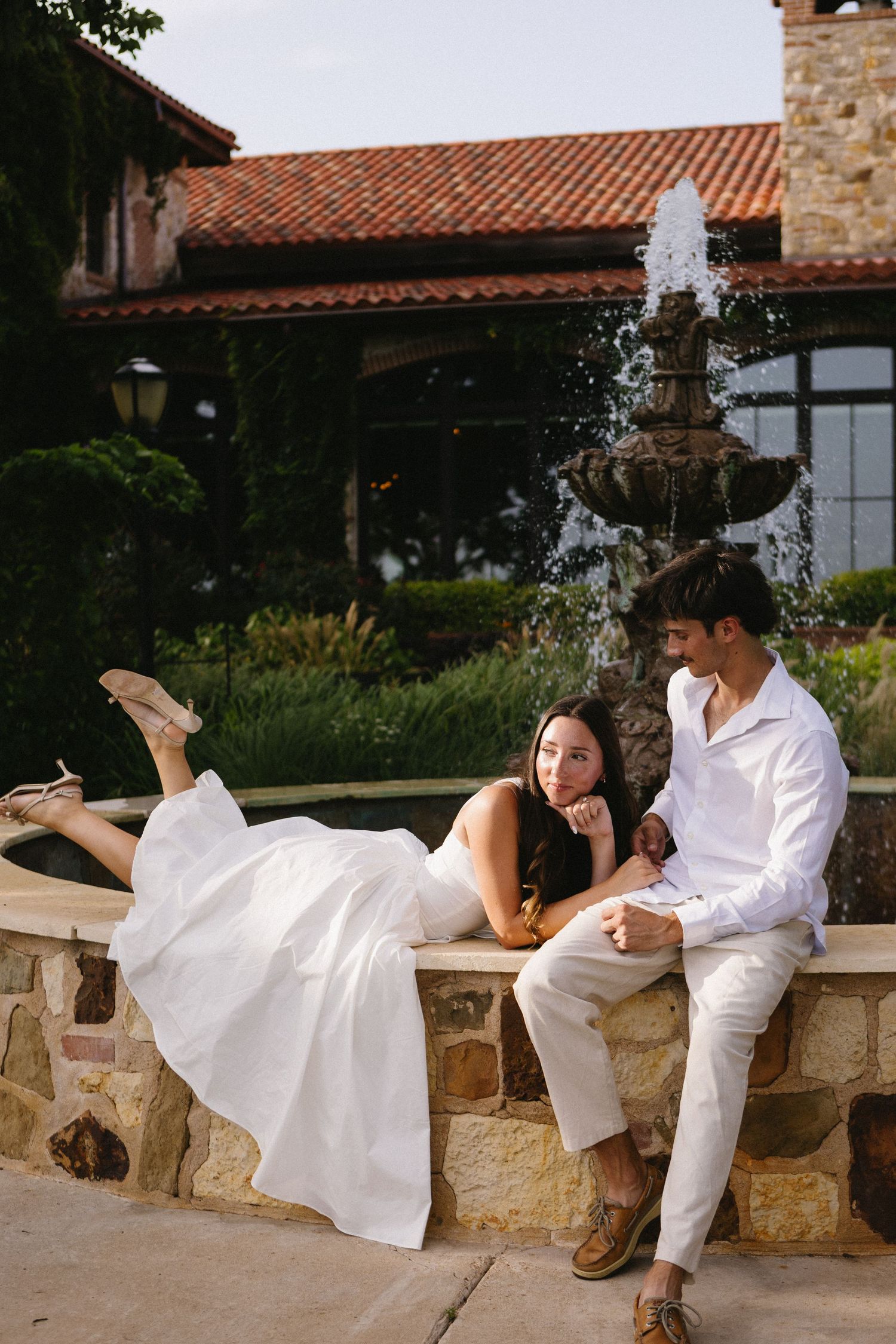 Couple in white attire sitting by a stone fountain outside a Spanish-style villa during golden hour.