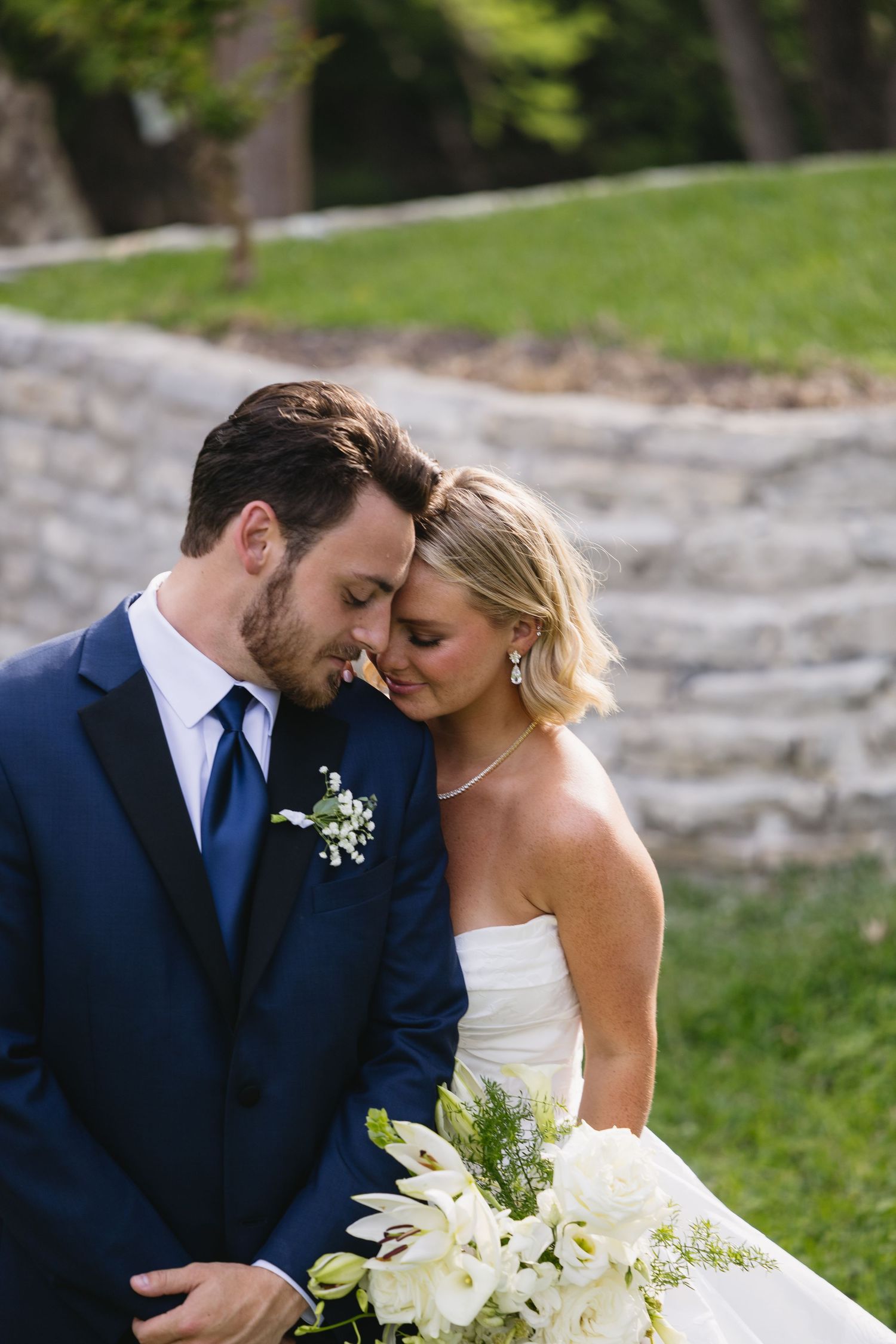 Bride and groom share an intimate moment outdoors, she holds white floral bouquet as he wears navy suit with boutonniere.