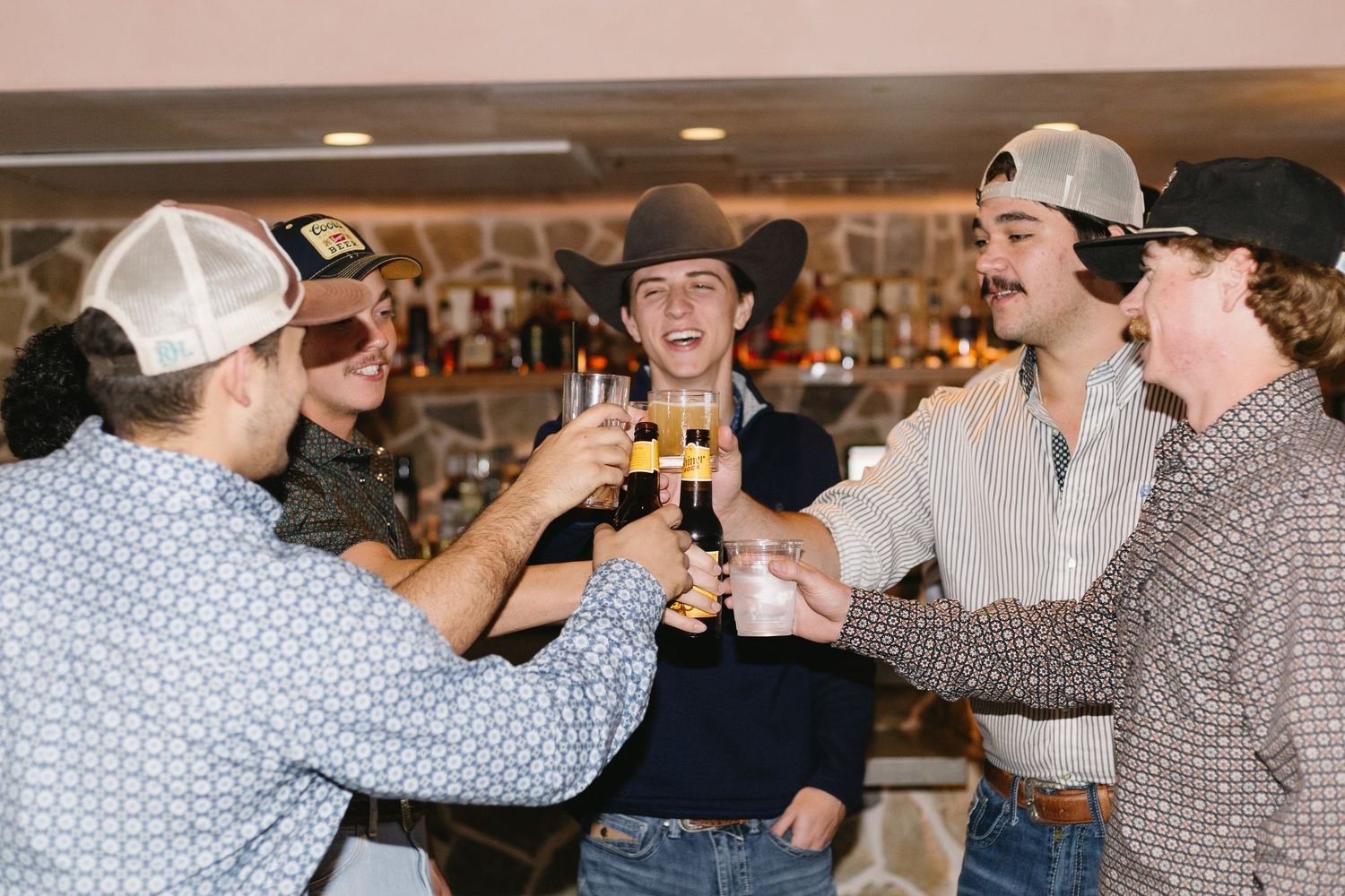 Group of young men in cowboy hats toasting drinks together at a bar, celebrating in western-style attire.