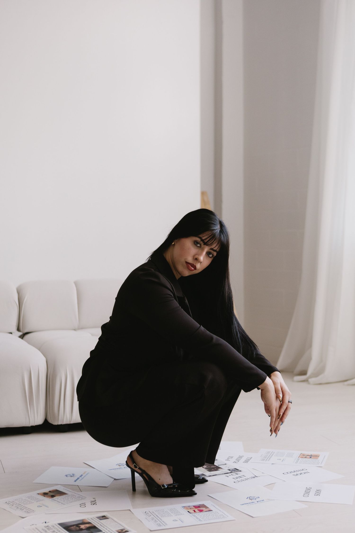 Woman in black outfit crouching on white floor surrounded by documents, with a white sofa in the background.